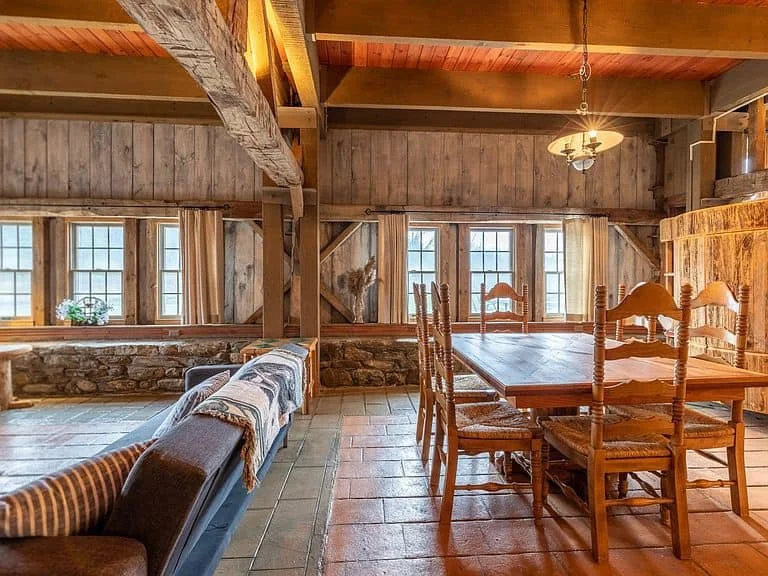 Rustic dining room with wooden beams, a long wooden table, and six wooden chairs, illuminated by a hanging chandelier, with multiple windows and exposed brick and wood wall decor.