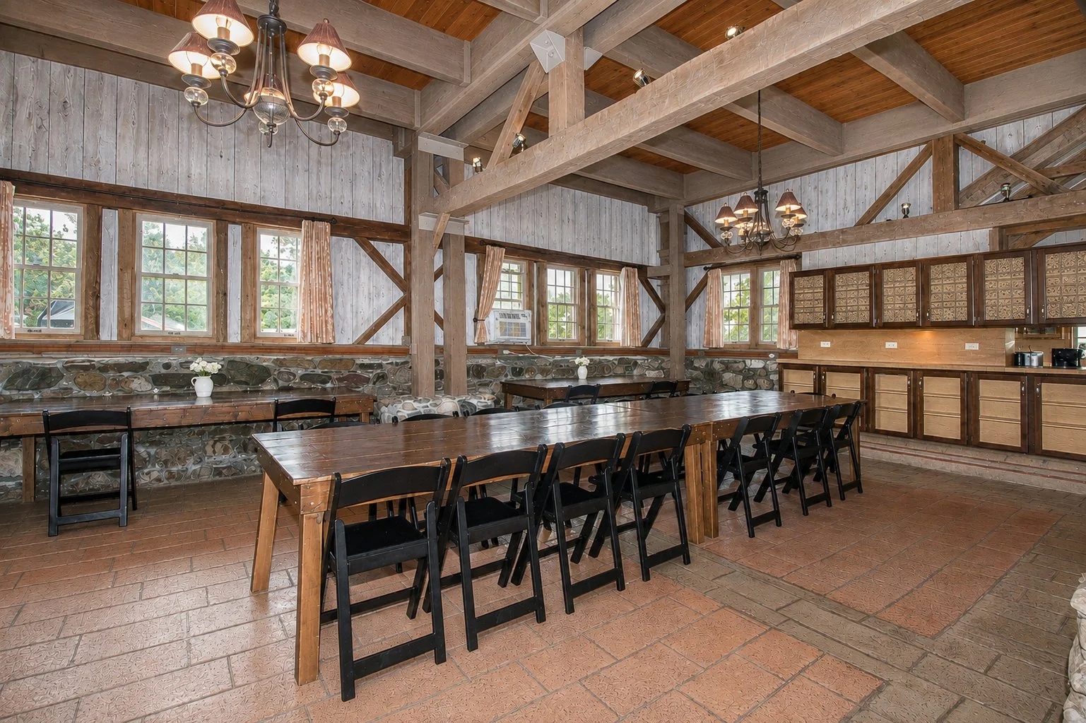 Interior of a rustic dining area with wooden beams, multiple windows with curtains, a long wooden table with black chairs, and a kitchen area with cabinets and a countertop in the background.