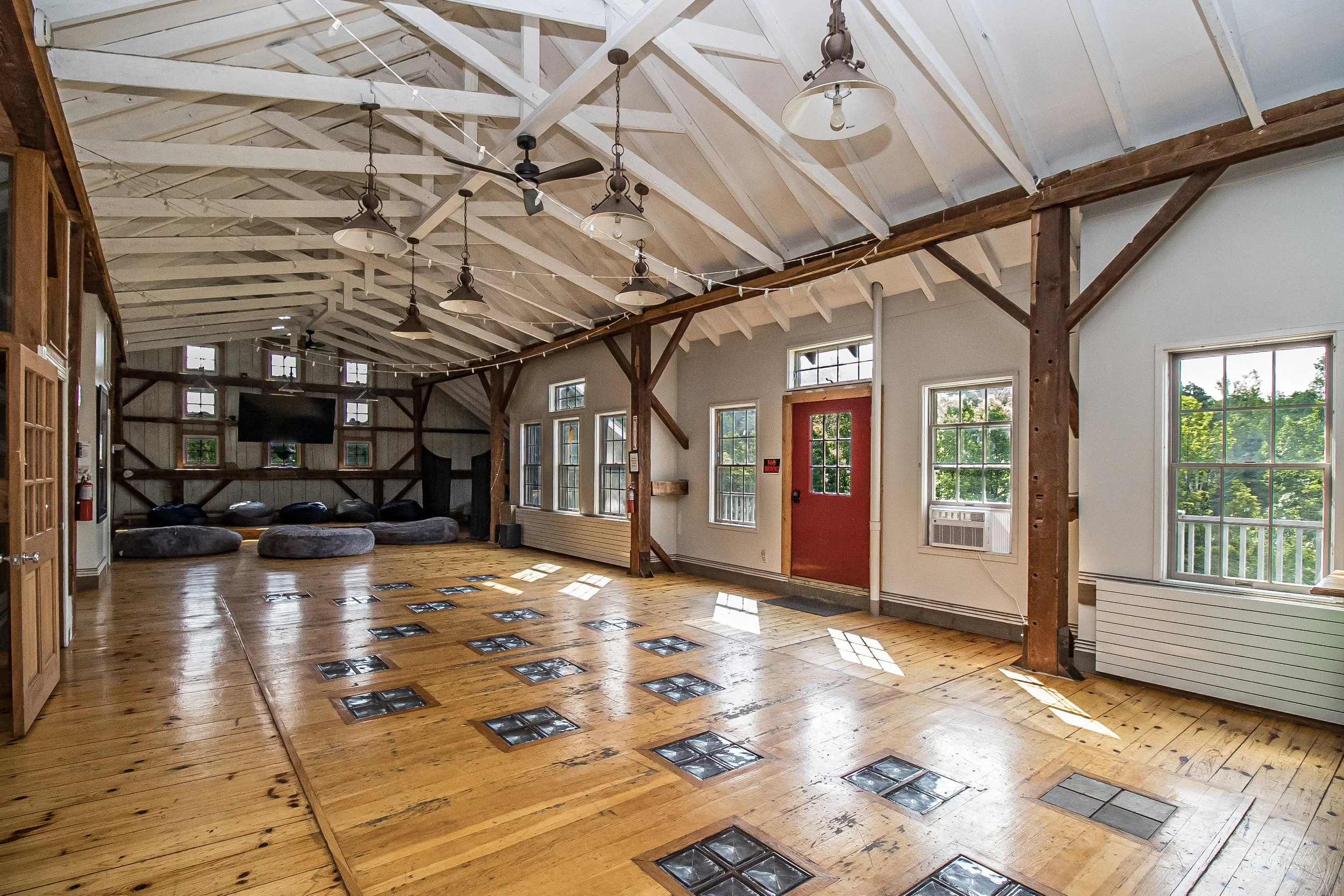 Interior of a spacious room with a high vaulted ceiling with exposed beams, multiple windows allowing natural light, wood flooring with glass blocks, and hanging light fixtures.