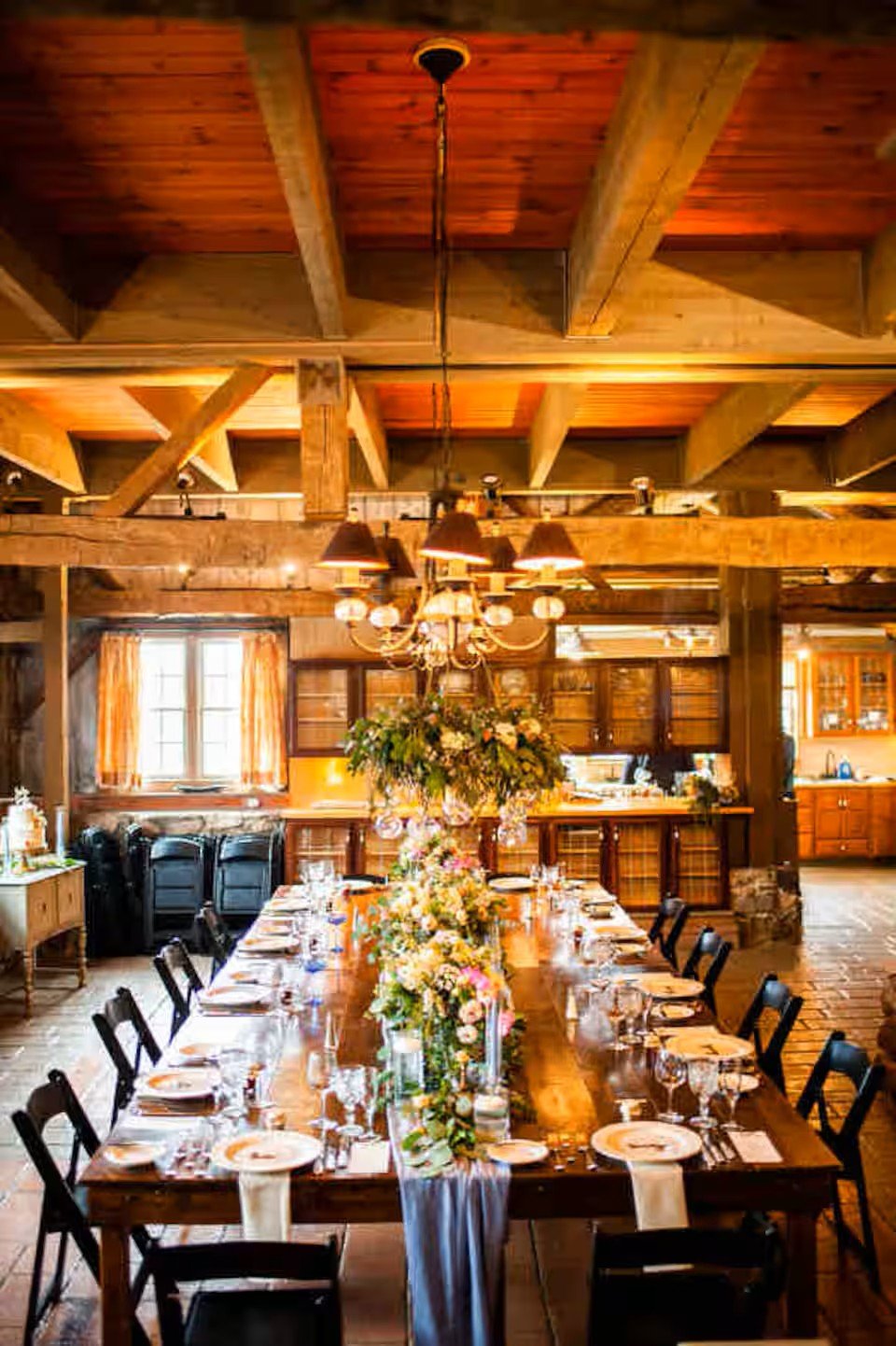 A rustic wooden dining room with a long table decorated with floral centerpieces, set for a meal with plates, glasses, and silverware, illuminated by a chandelier and natural light from windows.