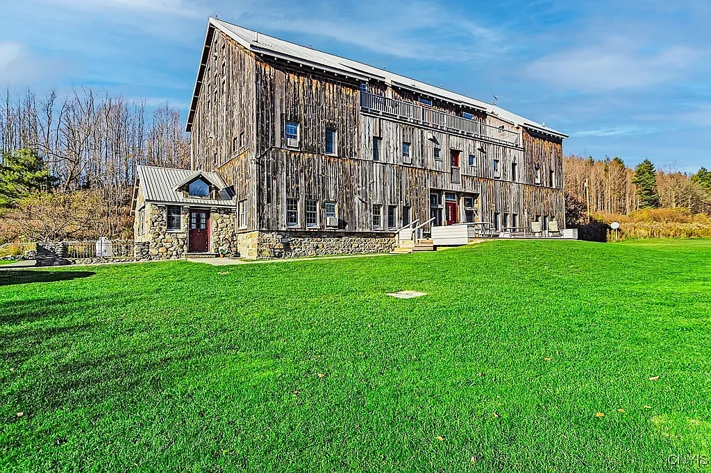A rustic wooden barn with a stone foundation and small side entrance, set on a large grassy lawn with trees in the background and a partly cloudy blue sky.