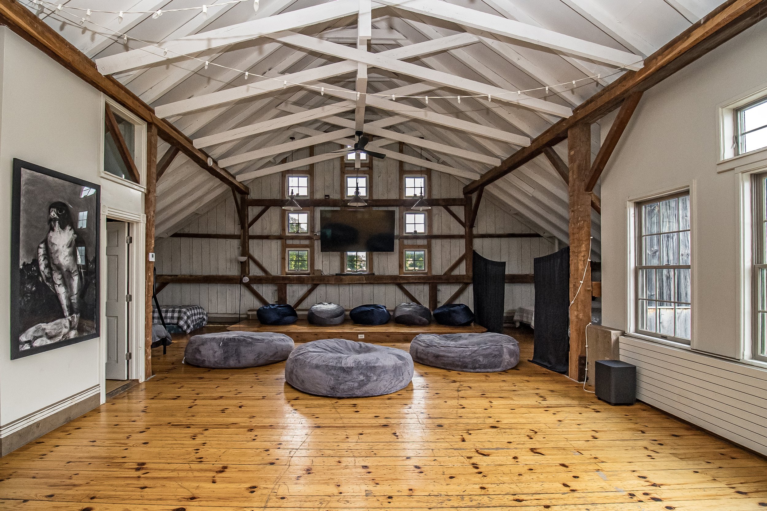 Interior view of a spacious farmhouse-style living room with exposed wooden beams, multiple windows, a large flat-screen TV, a ceiling fan, and a gathering of bean bag chairs on a wooden floor.