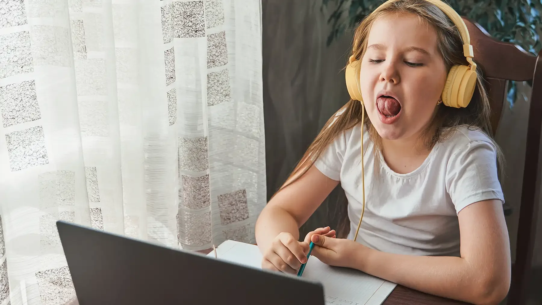 A young girl with long hair wearing a white shirt, large yellow headphones, and playing a musical instrument with her tongue stuck out at a laptop.