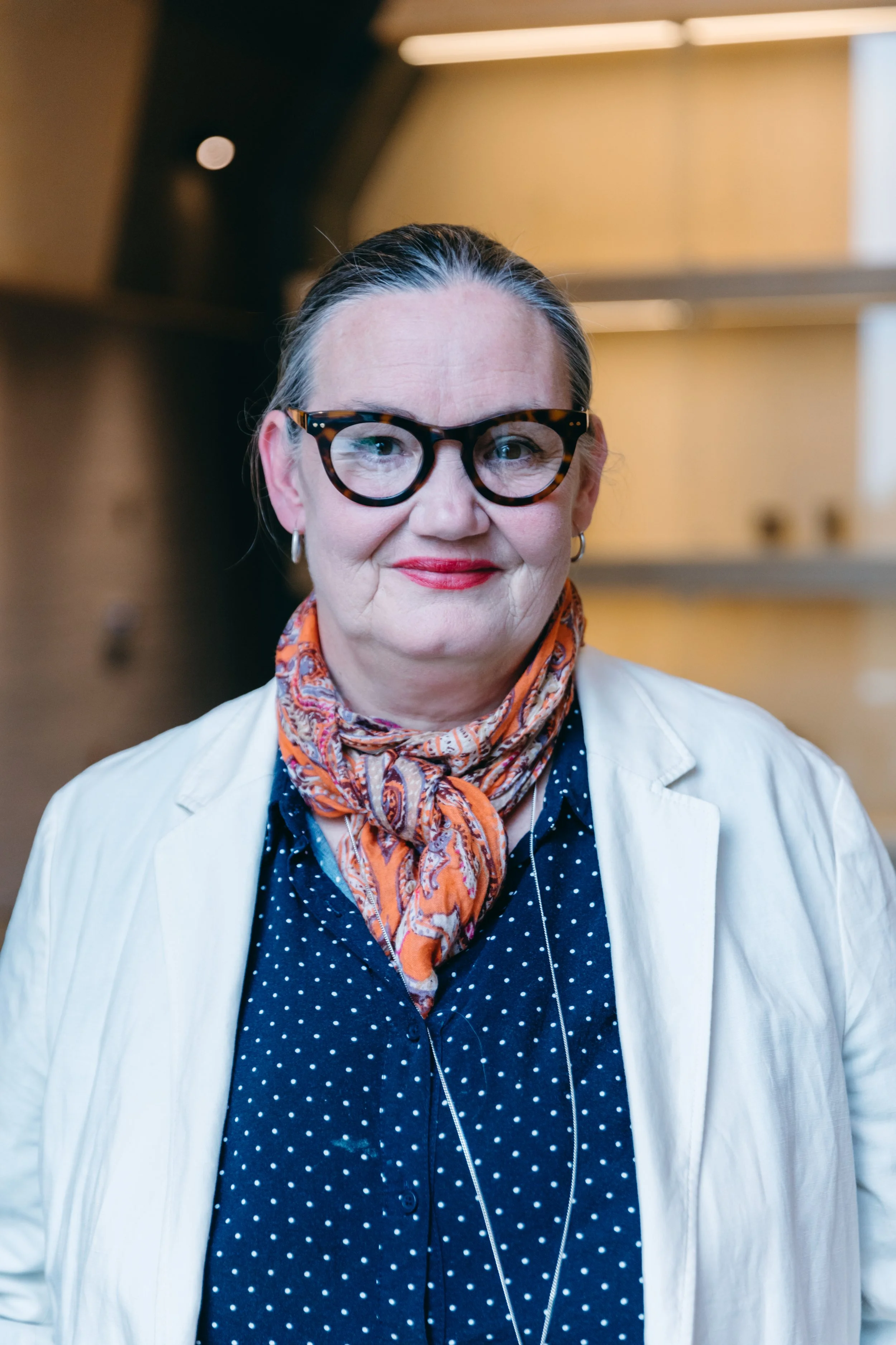 A woman with black and gray hair, wearing tortoiseshell glasses, red lipstick, a polka-dot navy blue shirt, an orange patterned scarf, a white blazer, and hoop earrings, smiling indoors.
