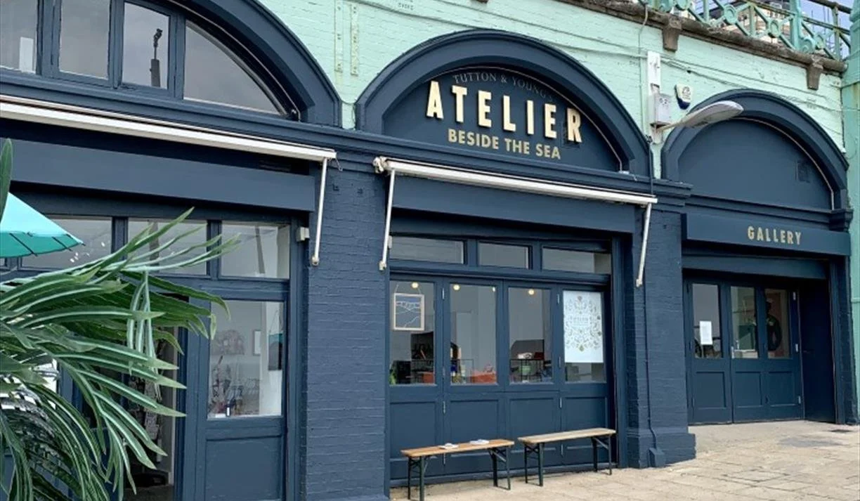 Exterior of an art gallery called Atelier beside the sea, with a blue facade and large arched windows, a small bench outside, and potted plants nearby.