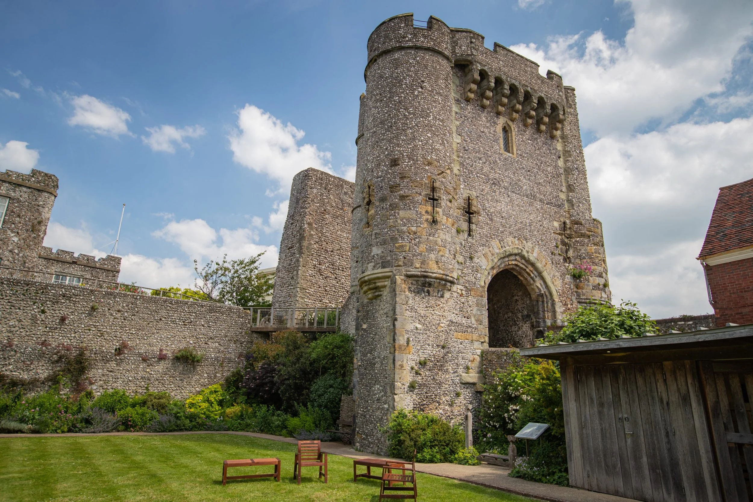 Historic stone castle with a round tower and battlements, surrounded by a garden with benches, trees, and a wooden shed, under a partly cloudy sky.