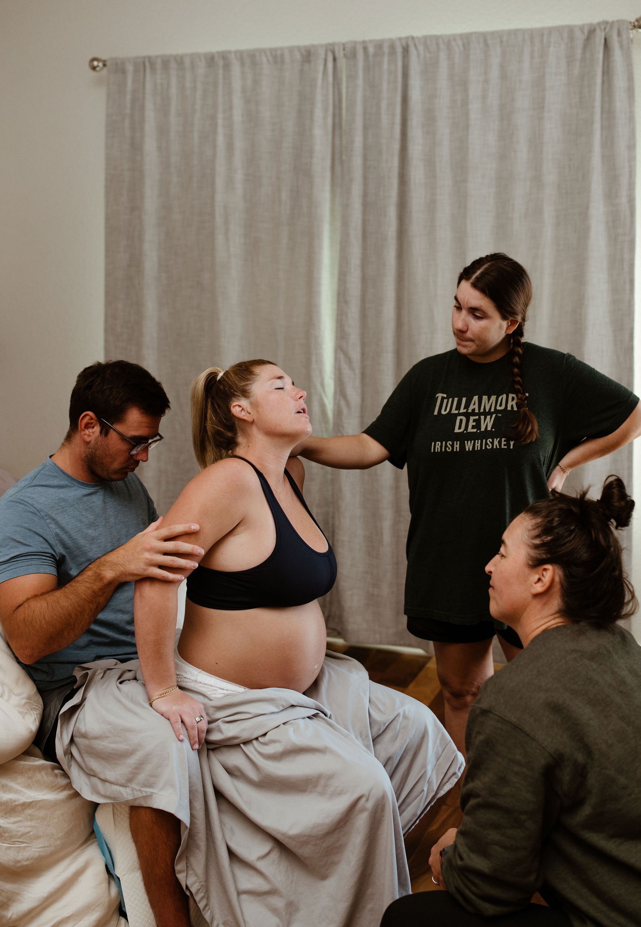 A group of five people, four women and one man, gathered around a pregnant woman sitting on a bed, receiving emotional support, with curtains in the background.