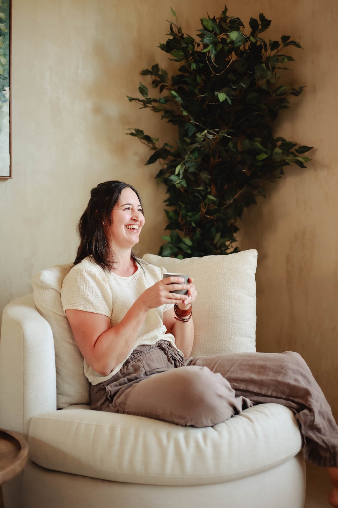 A woman sitting on a cream-colored sofa, smiling and holding a mug, with a large green leafy plant behind her and beige walls.