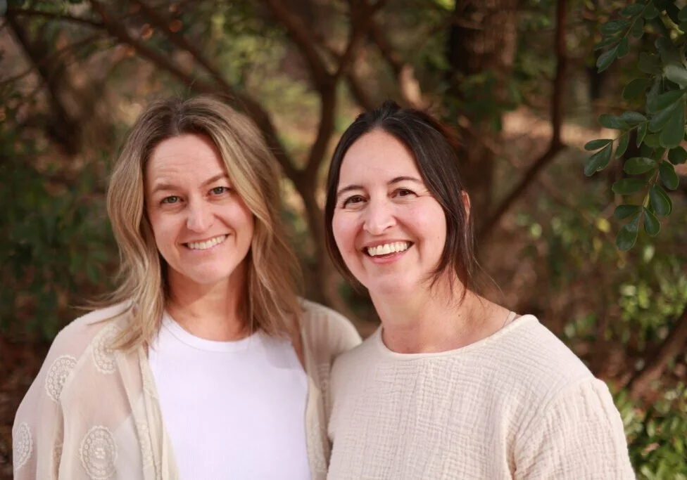 Two women smiling outdoors in front of trees and foliage.