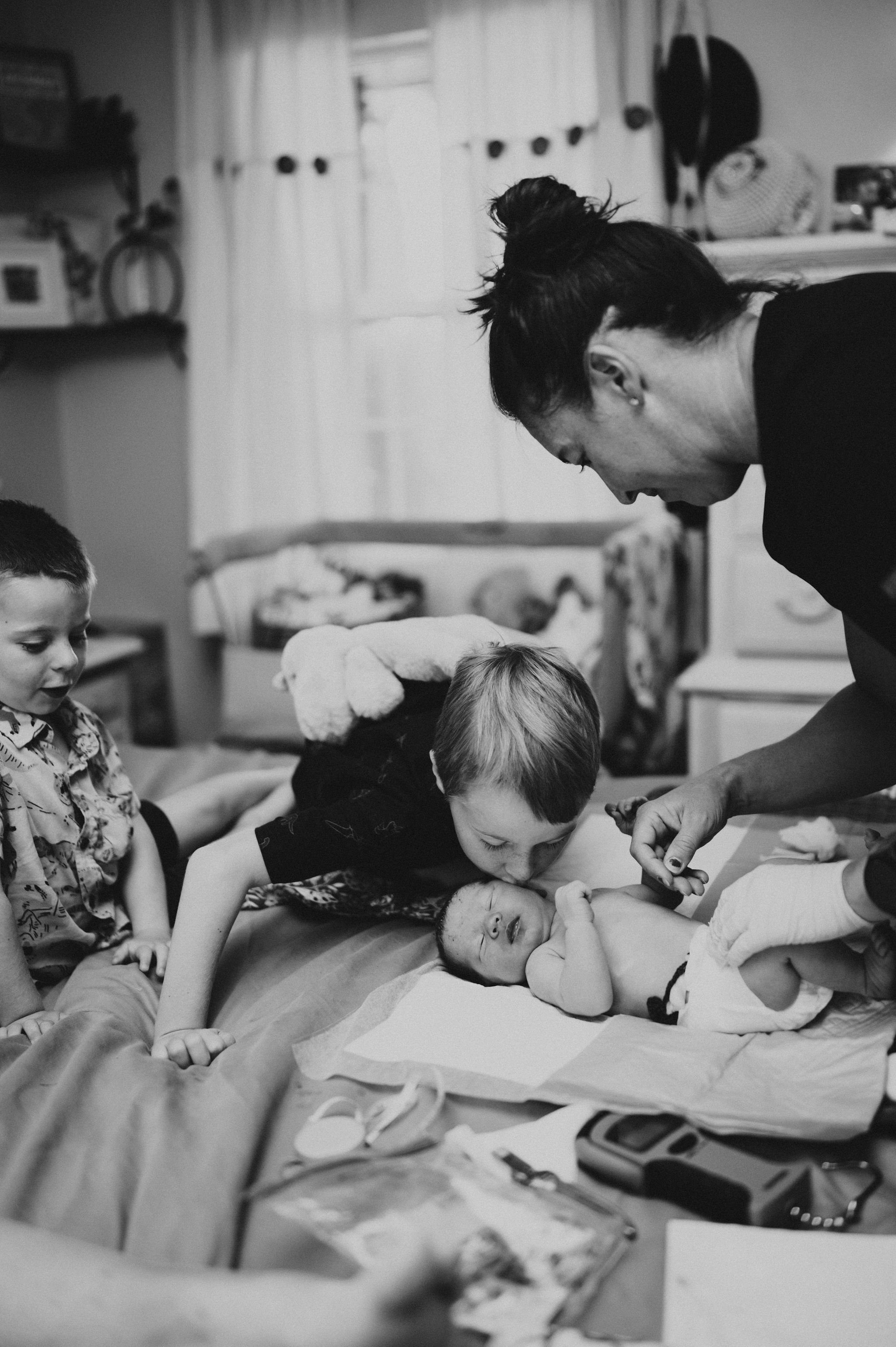 A black and white photo showing a woman and children gathered around a newborn baby lying on a hospital bed, possibly during a home birth or similar setting. The woman is kissing the baby, and the children are observing closely. Medical tools and supplies are visible on the table nearby.