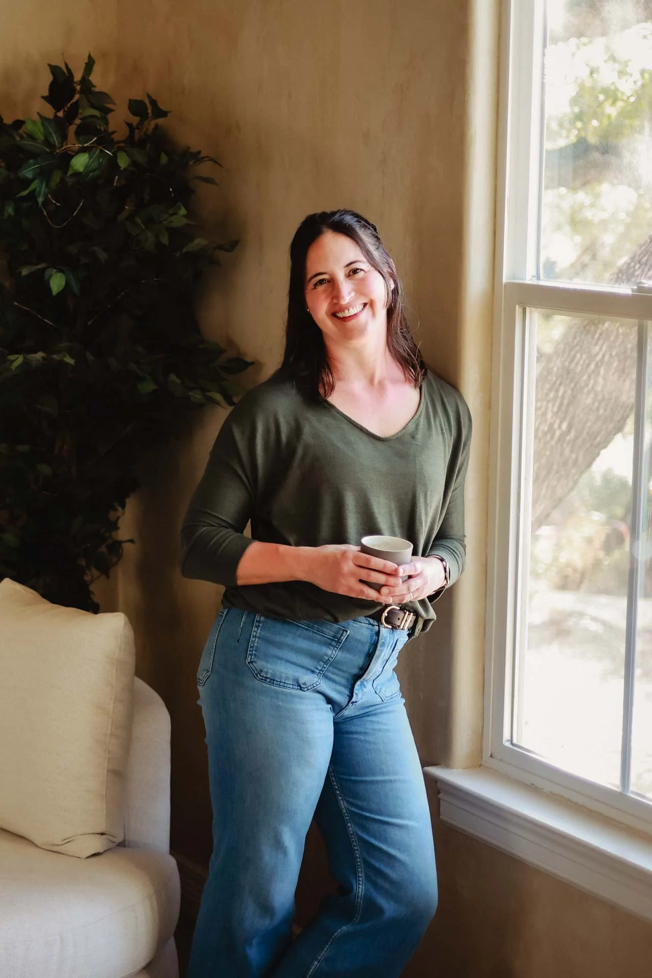 A woman with dark hair, wearing a green long-sleeve shirt and blue jeans, standing indoors by a window, smiling with a cup in her hands.