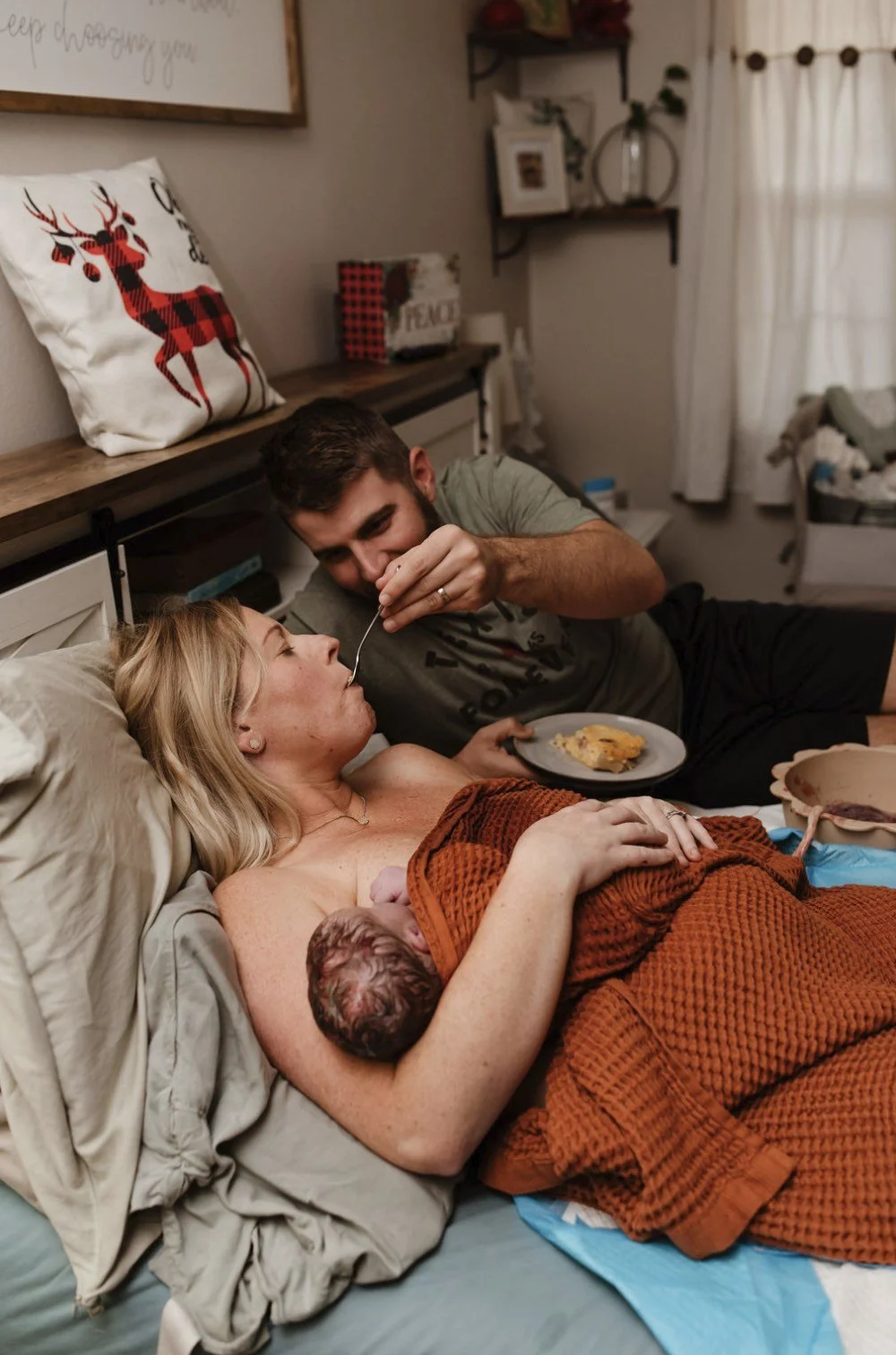 A woman is lying in bed after giving birth, with a newborn baby resting on her chest. A man is feeding her with a spoon while she looks at him. The setting is cozy with a decorated pillow and shelves in the background.