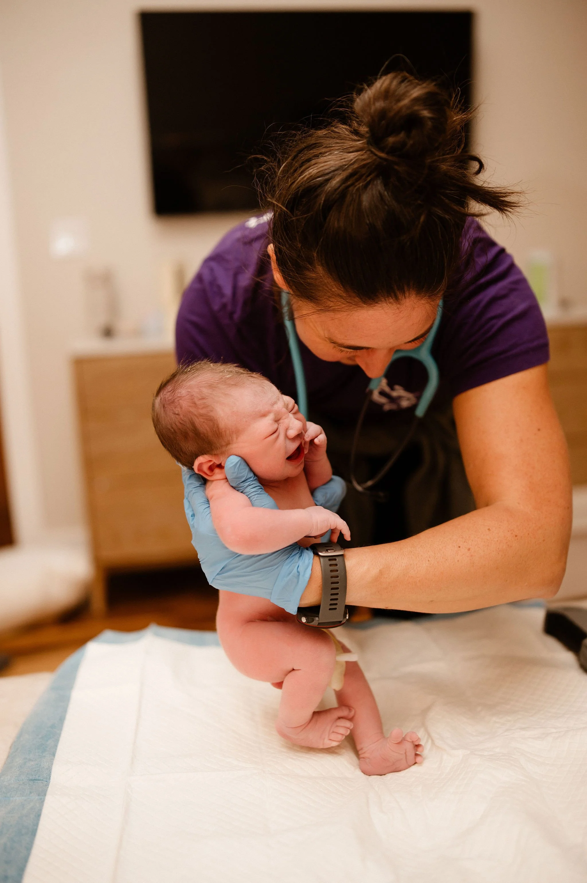A healthcare professional, wearing purple scrubs and gloves, is holding and comforting a crying newborn baby on a changing pad.