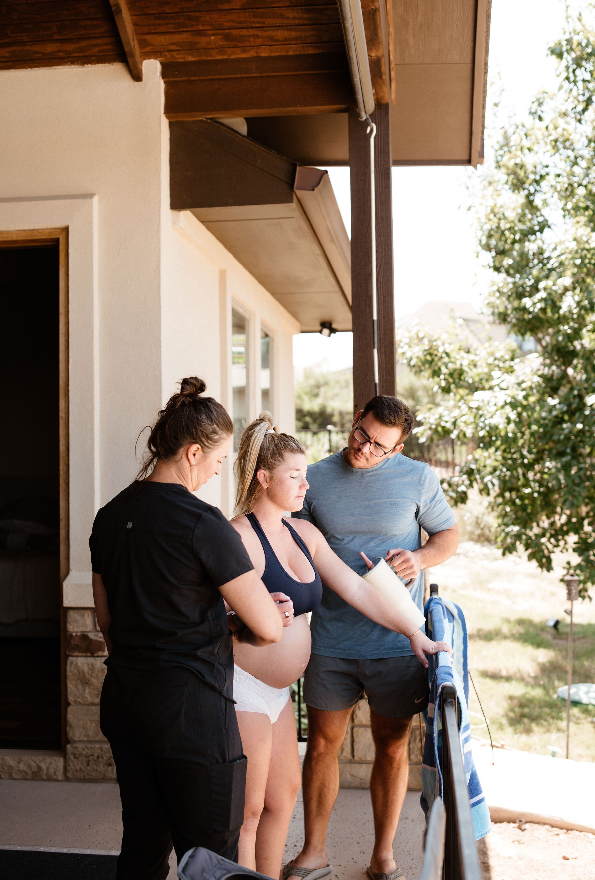 A group of four people standing outside a house, including a pregnant woman, with three of them looking at a tablet and engaging in conversation. The setting is a sunny day with trees in the background.