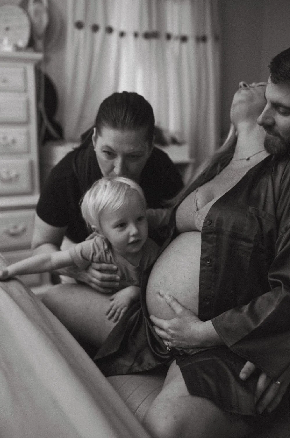 A woman giving birth in a hospital room, with a young child and two adults present, one of whom is supporting the woman and the other observing.