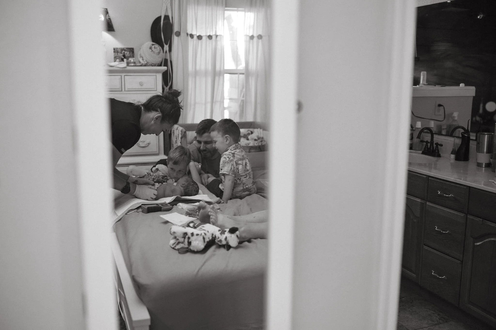 Black and white photo of a woman and children in a bedroom, gathered around a bed, with a crib and dresser visible, viewed through a partially open door.