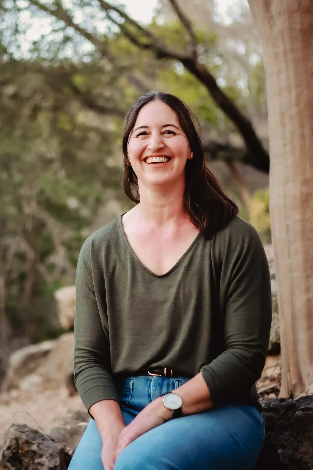 A woman with shoulder-length dark hair, wearing a green long-sleeve shirt, blue jeans, and a wristwatch, sitting outdoors on a rock near a tree, smiling at the camera.