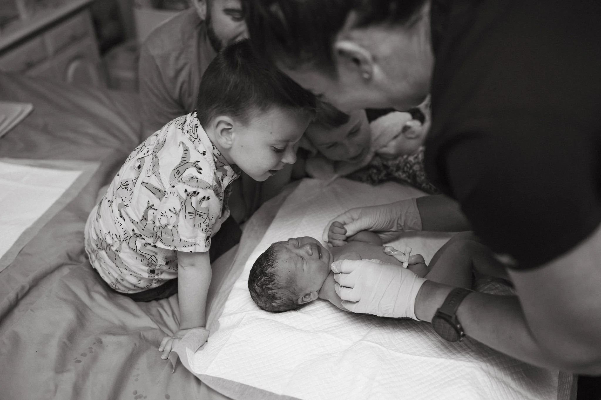 A baby lies in a hospital bed with a bandage on their head, surrounded by three children and adults, who are gathered around, showing support and care during the birth process.