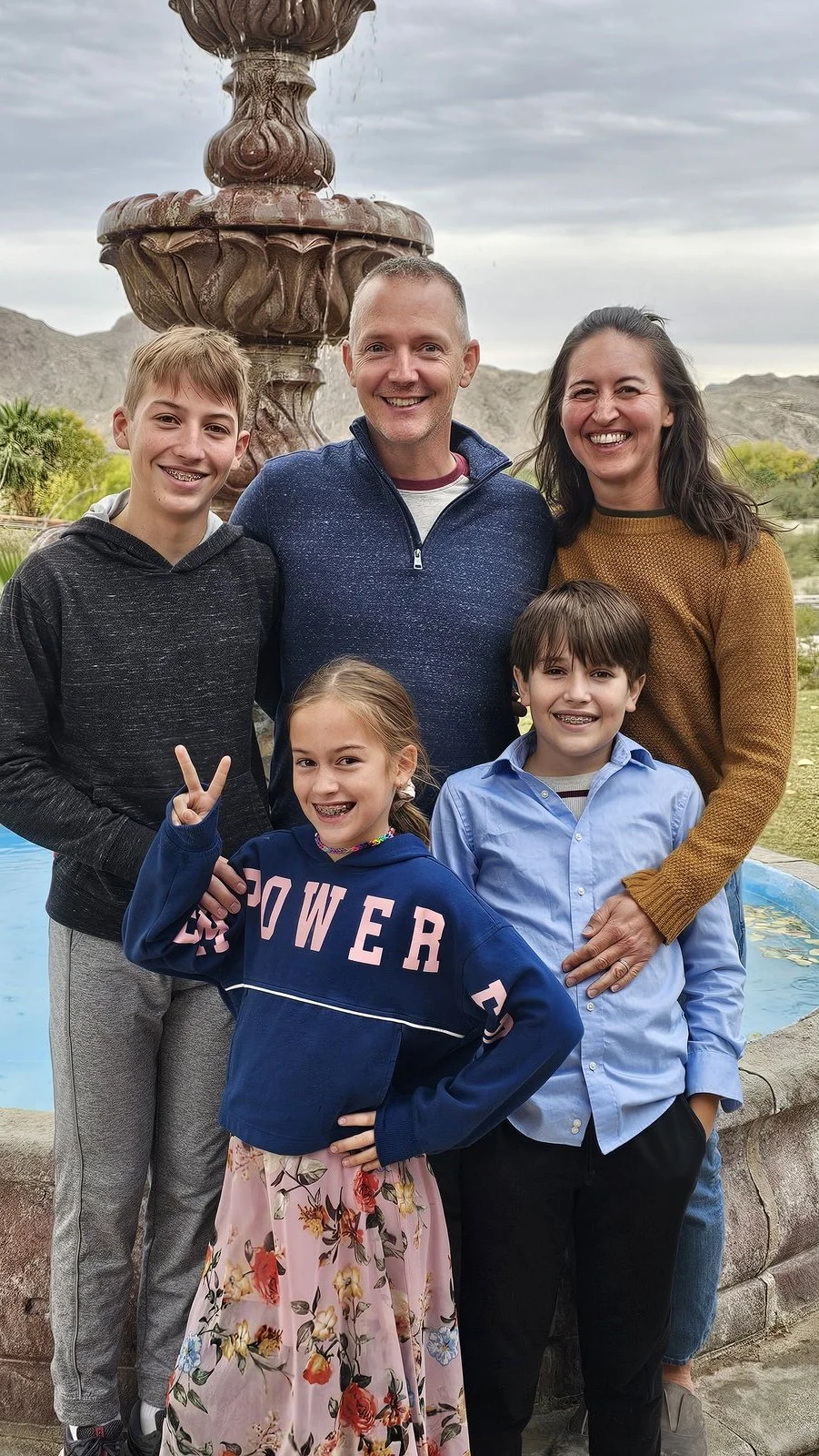 A family of six smiling and posing outdoors in front of a decorative fountain and desert landscape with mountains in the background.