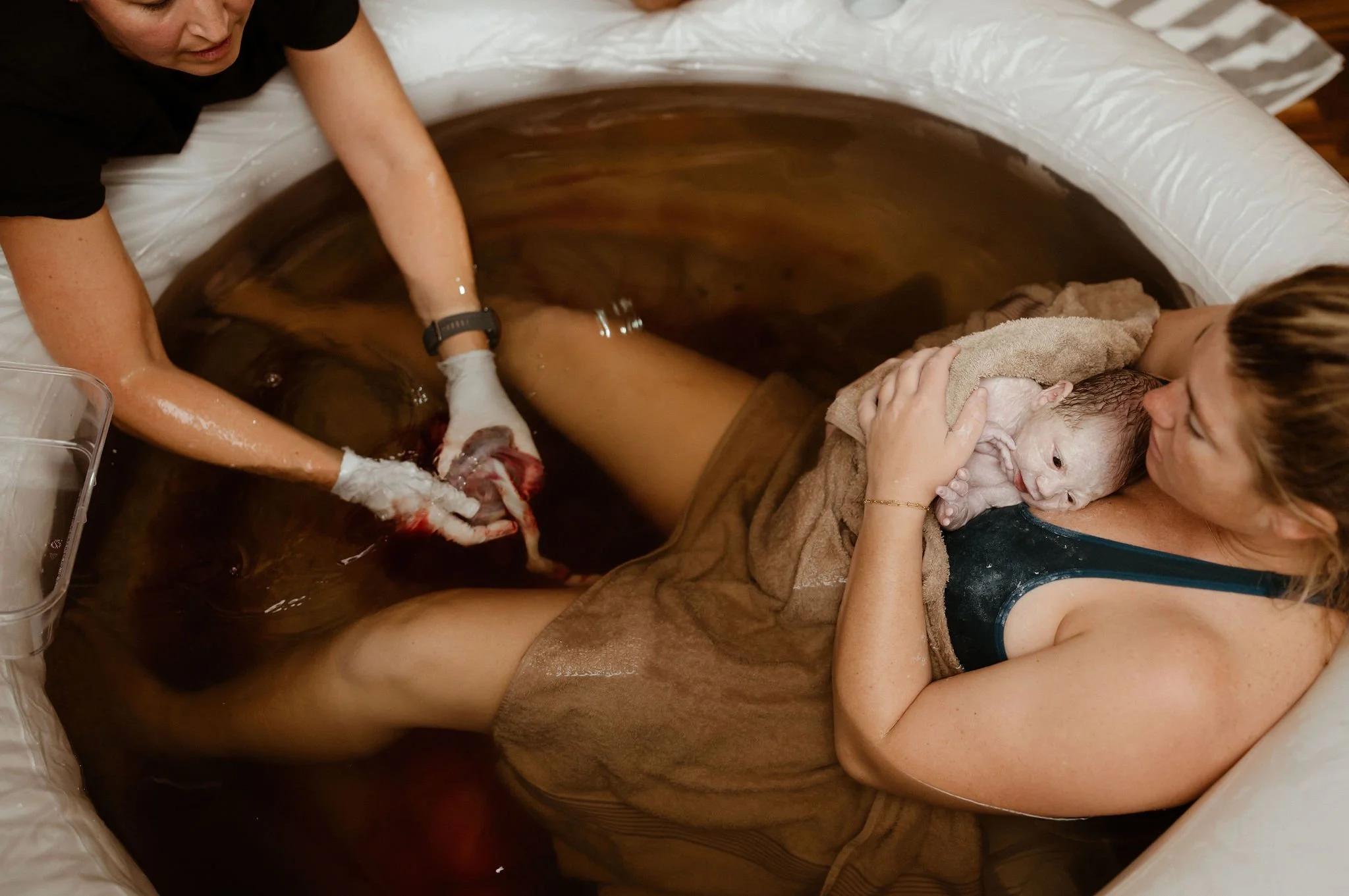 A woman is giving birth in a birthing pool, supported by another woman who is gently holding her and covering her face with a towel.