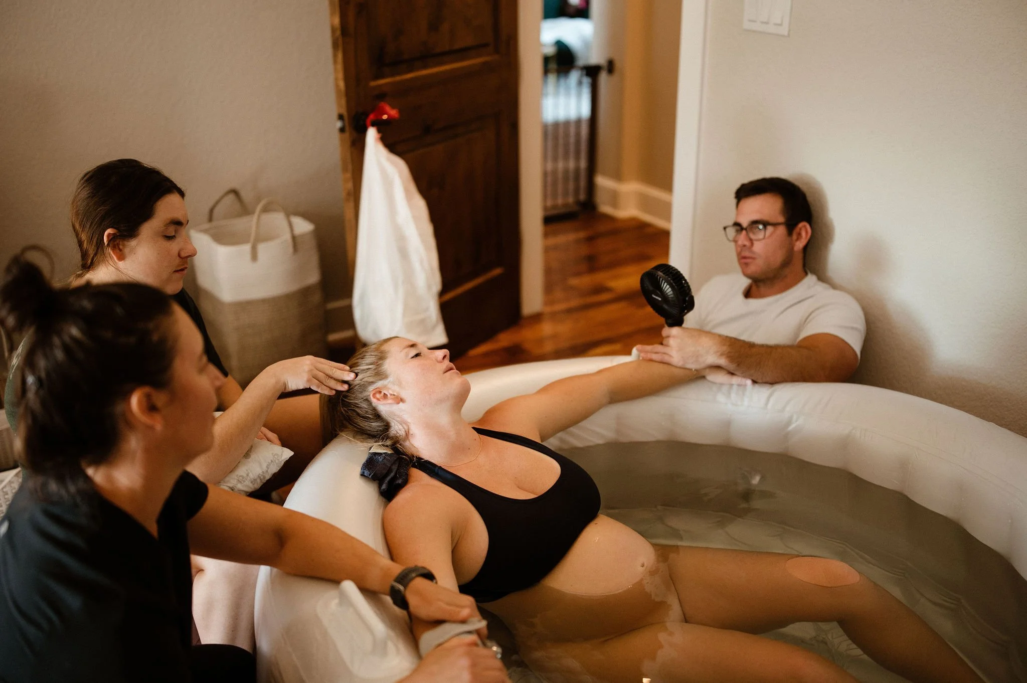 People gathered around a woman in a bathtub, some comforting her and others holding a handheld fan in a home setting.