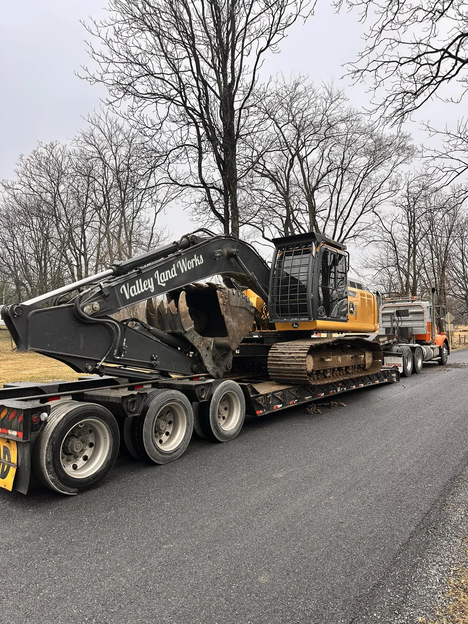 A large excavator labeled Valley Land Works is loaded onto a flatbed truck on a paved road, with leafless trees in the background under gray overcast sky.