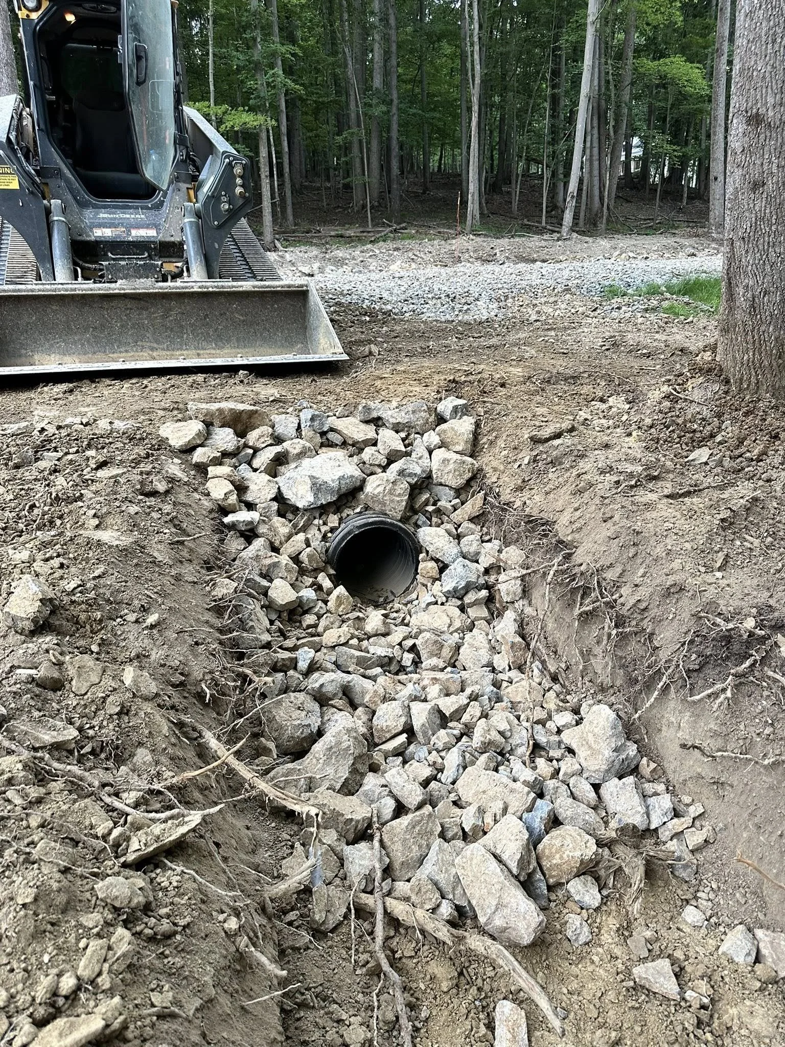 A construction site in a wooded area shows a large excavator with a bucket attachment in the background. The foreground features a drainage pipe surrounded by rocks and dirt, indicating ongoing drainage or piping work.