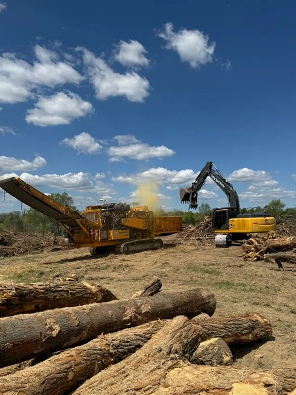 Construction site where logs are being sawed and chopped with heavy machinery under a partly cloudy sky.