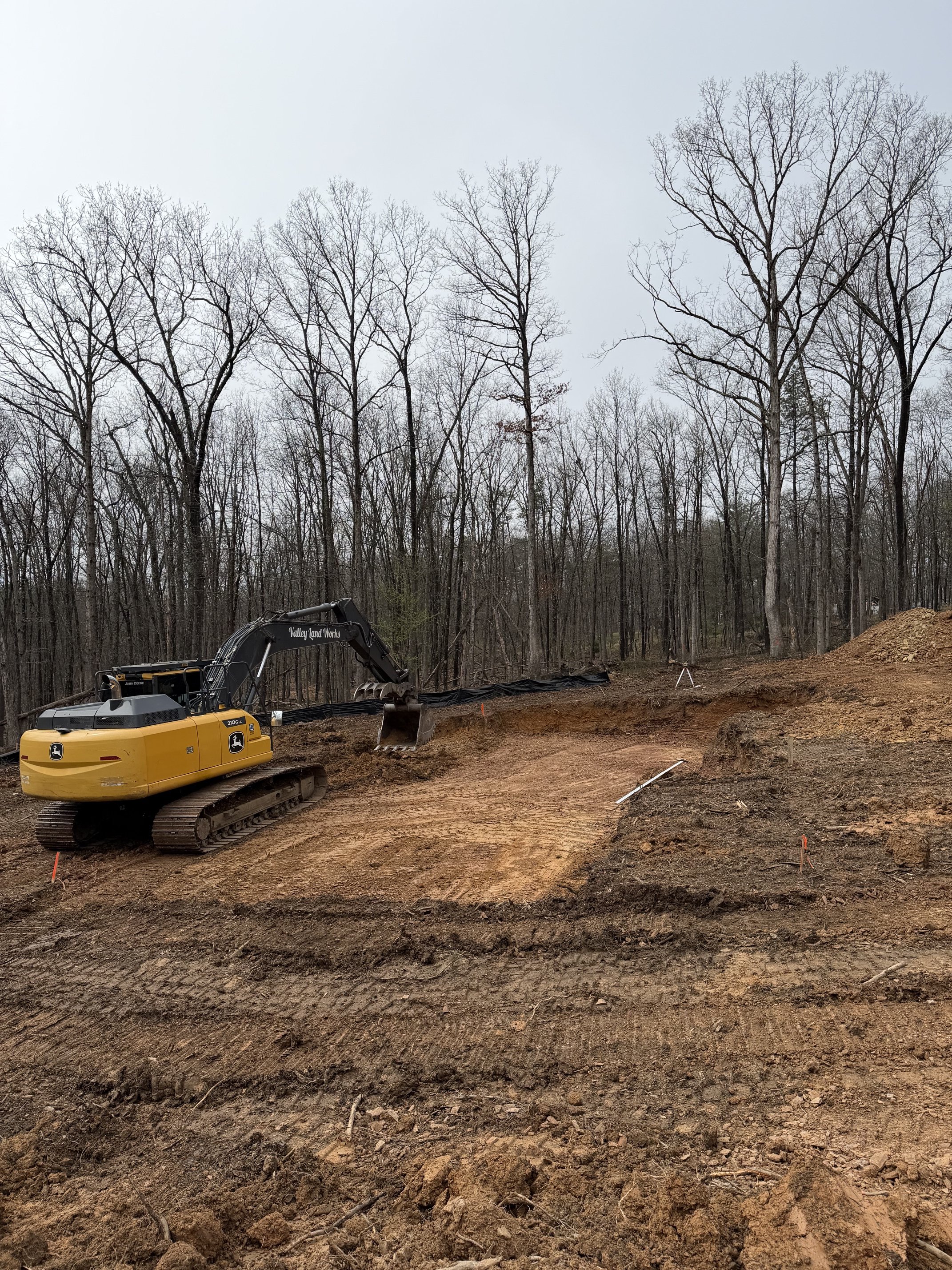 A construction site with excavated dirt ground and a yellow John Deere excavator. Bare trees with no leaves and a cloudy sky in the background.