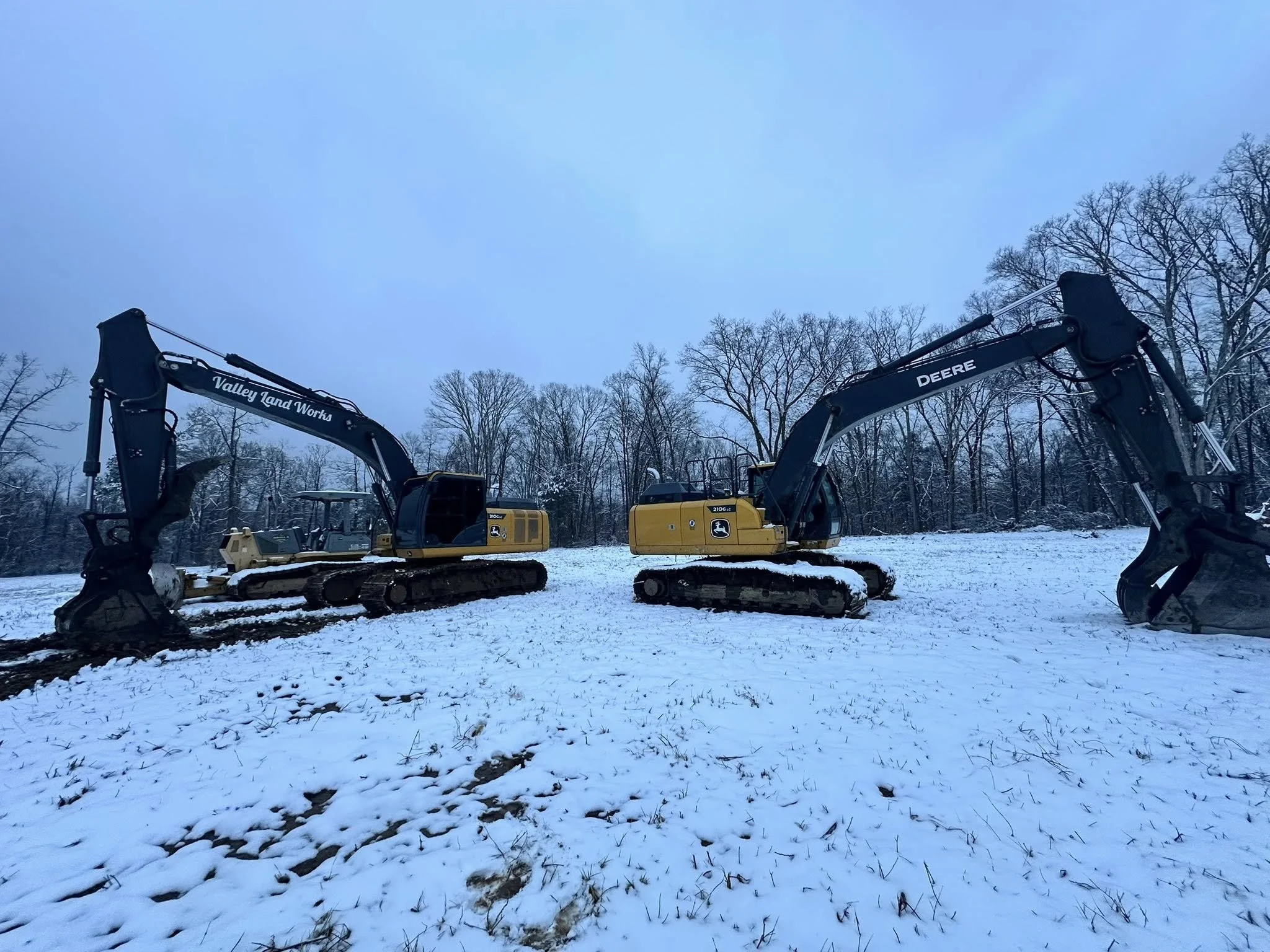 Several yellow and black John Deere excavators parked outside in a snowy area surrounded by leafless trees.