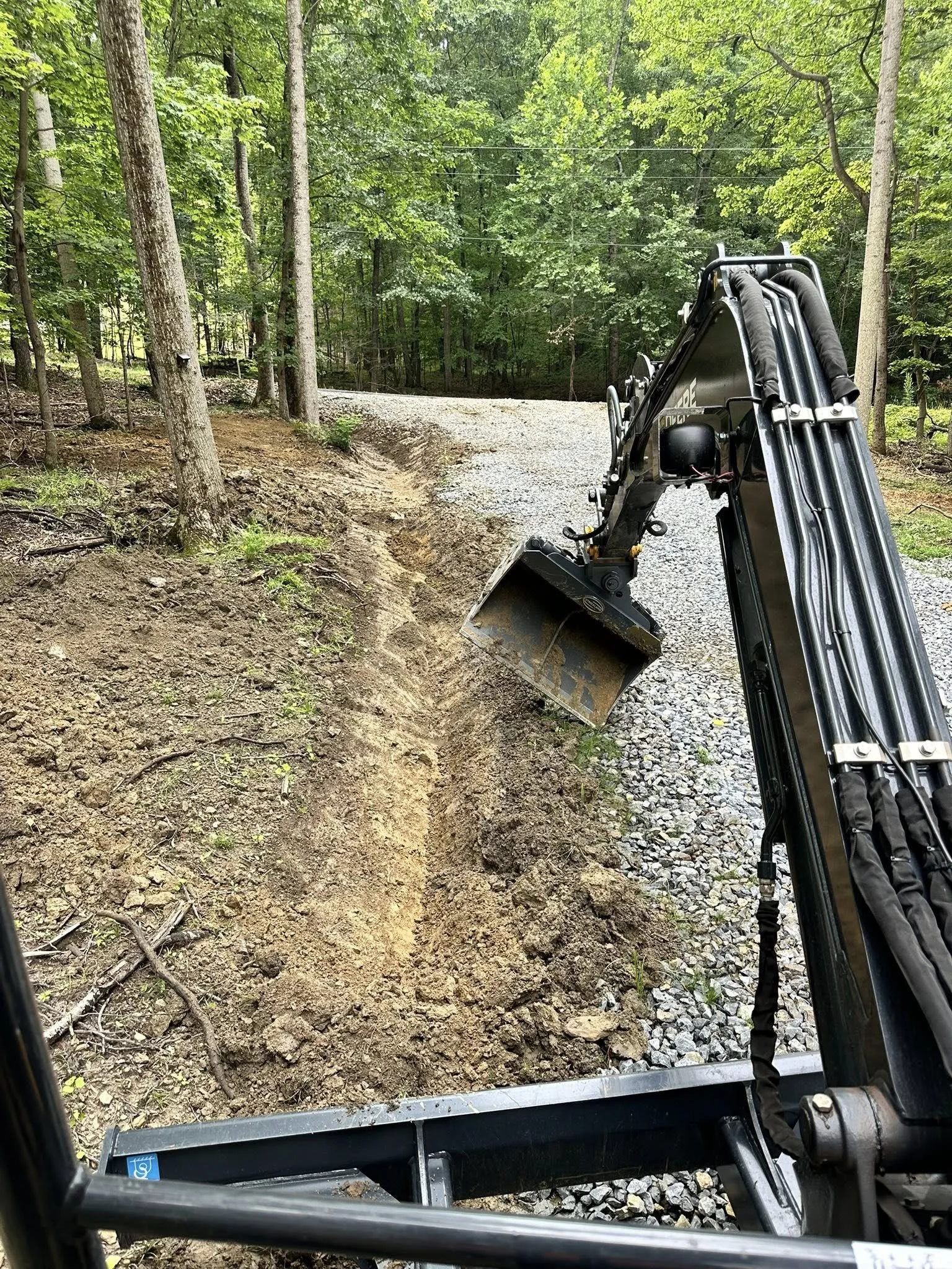 View from a construction vehicle showing a dirt and gravel trail in a dense green forest with trees on both sides.
