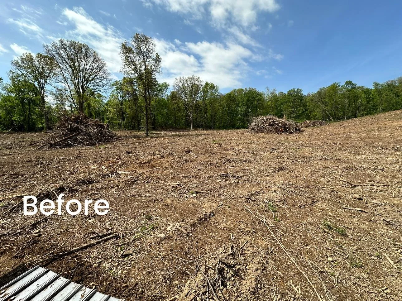 A cleared land area with dry, bare soil and scattered small branches. In the background, there are several piles of cut tree branches and a line of green trees under a partly cloudy blue sky.