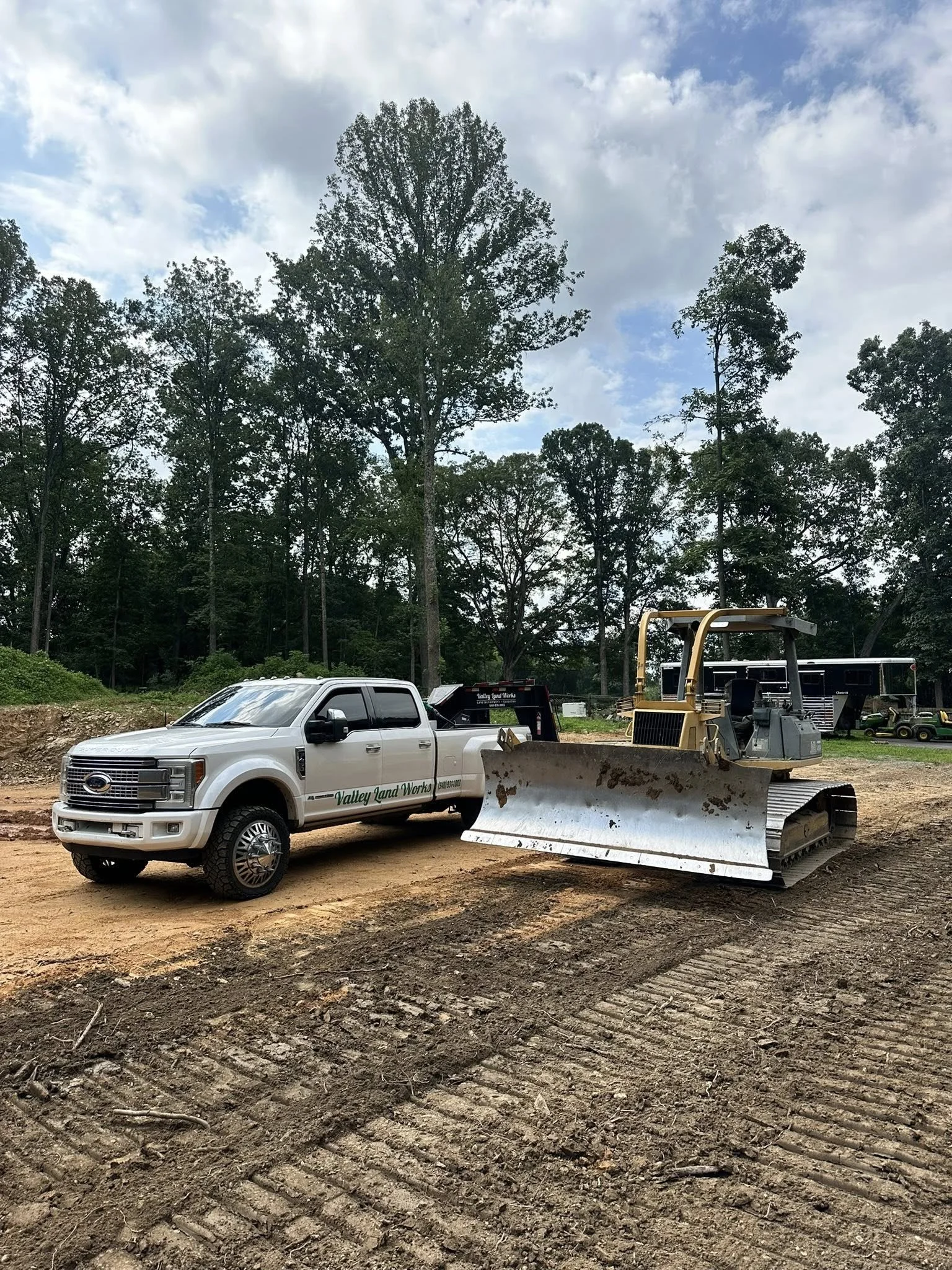 A white pickup truck labeled 'Valley Land Works' is parked on a muddy construction site. Beside it is a large yellow and gray bulldozer. In the background, tall trees with dense green foliage are under a partly cloudy sky.
