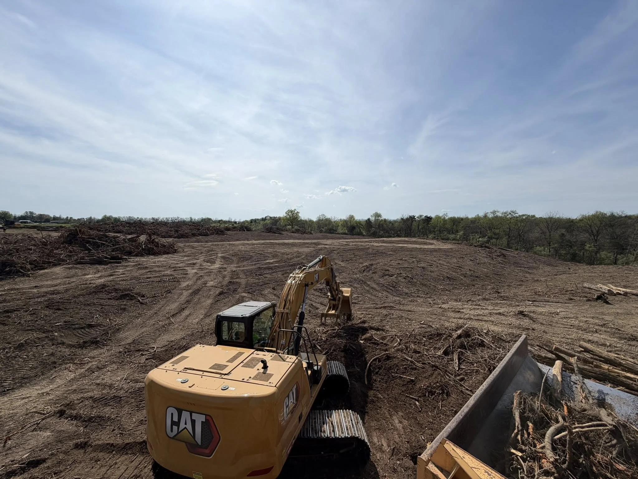 Construction site with earth-moving equipment and cleared land, trees in the background, under a partly cloudy sky.