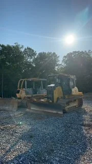 Two bulldozers parked on gravel under a sunny sky with trees in the background.