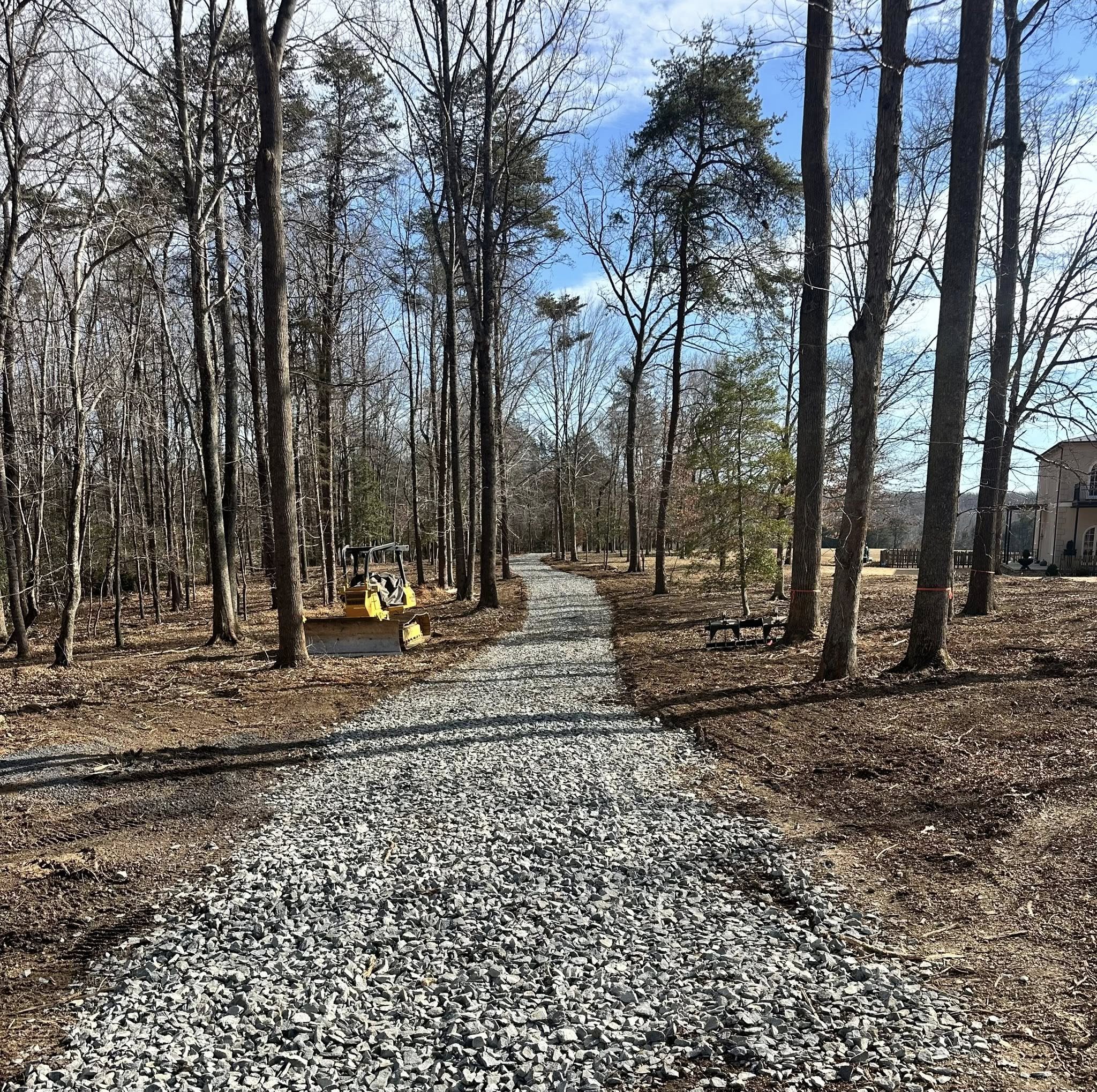 A wooded backyard with bare trees, a gravel pathway, and a yellow lawn mower parked to the left, under a partly cloudy sky.
