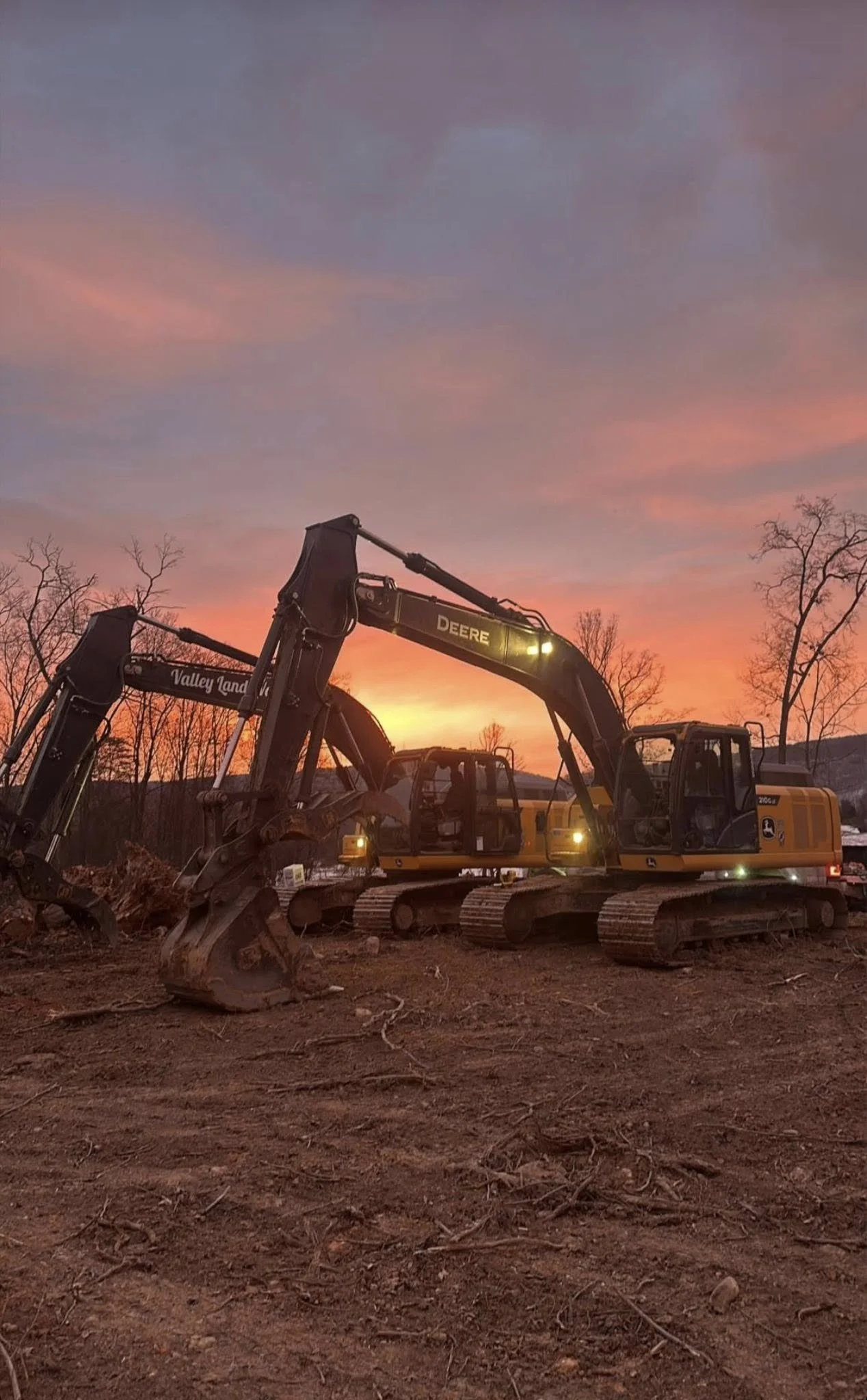 Two large yellow and black excavators parked on dirt ground during sunset, with a colorful sky and leafless trees in the background.