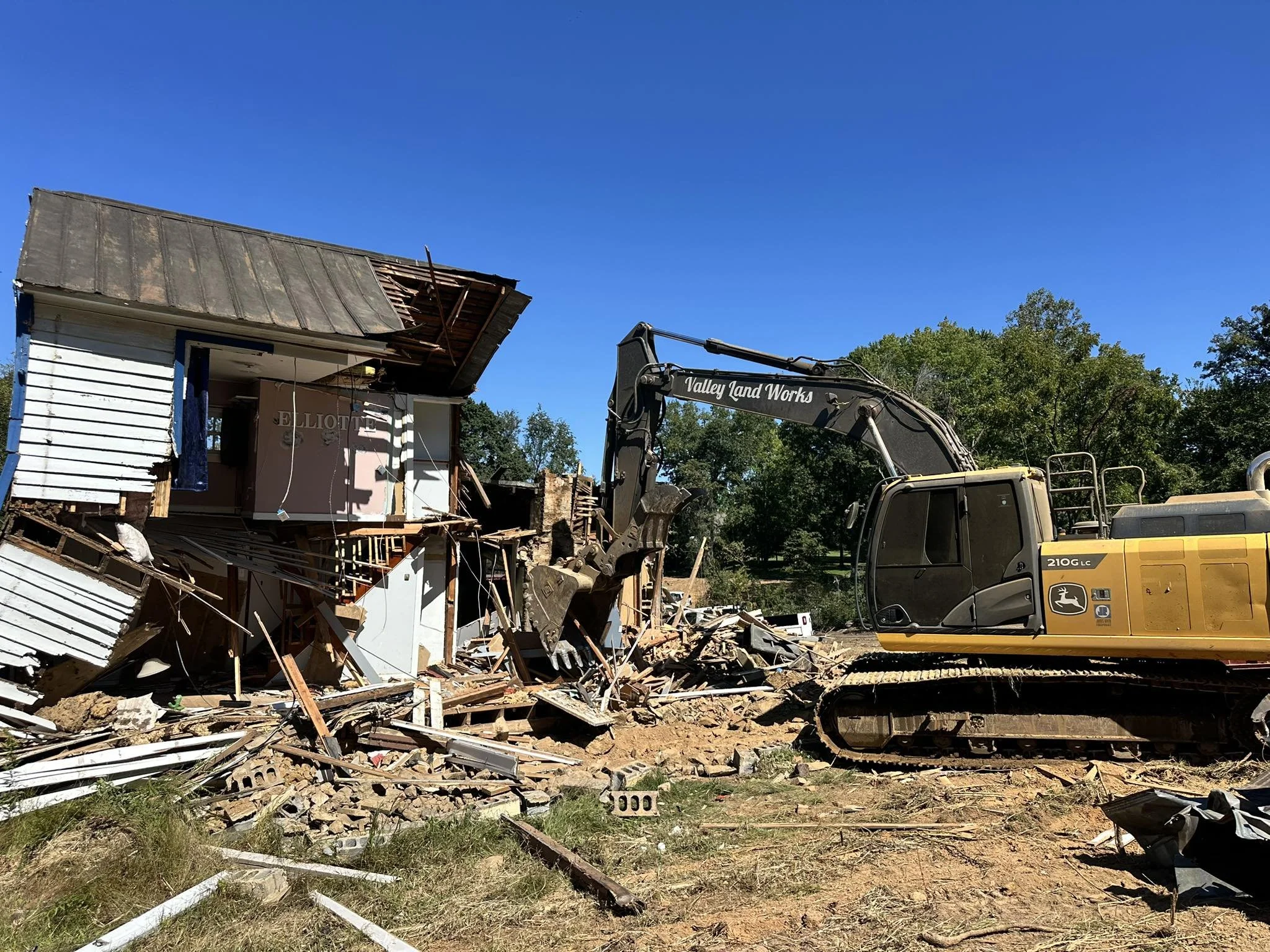 A construction excavator tearing down a house in a rural area under a clear blue sky.