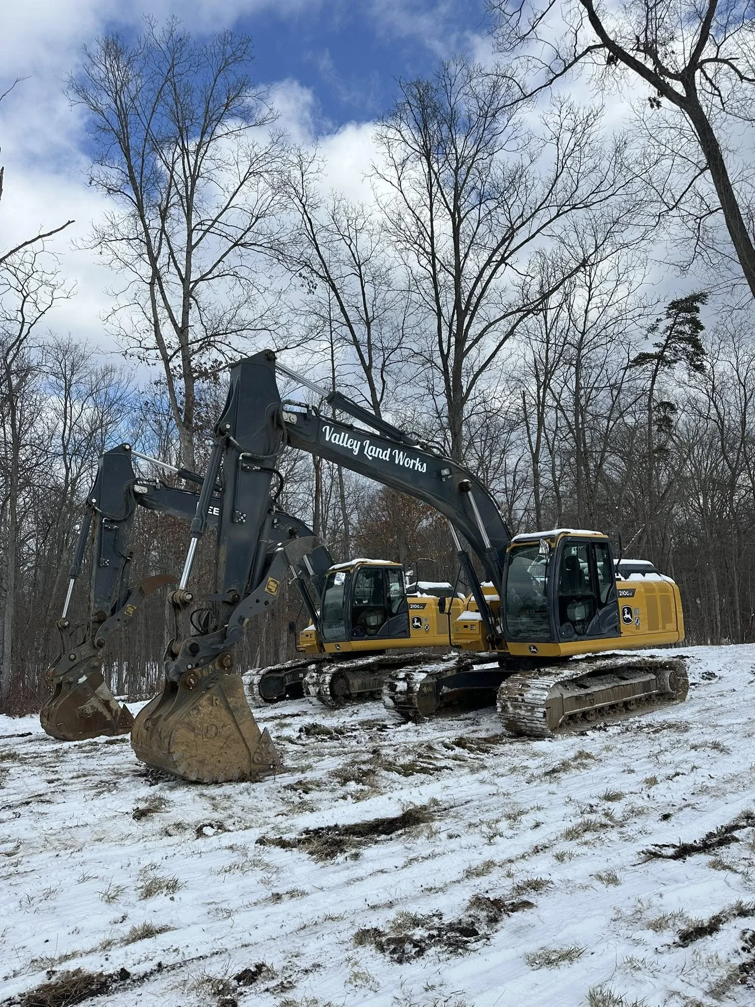 A yellow and black excavator with the label 'Valley Land Works' on its arm, parked on snow-covered ground with patches of grass, surrounded by leafless trees against a partly cloudy sky.