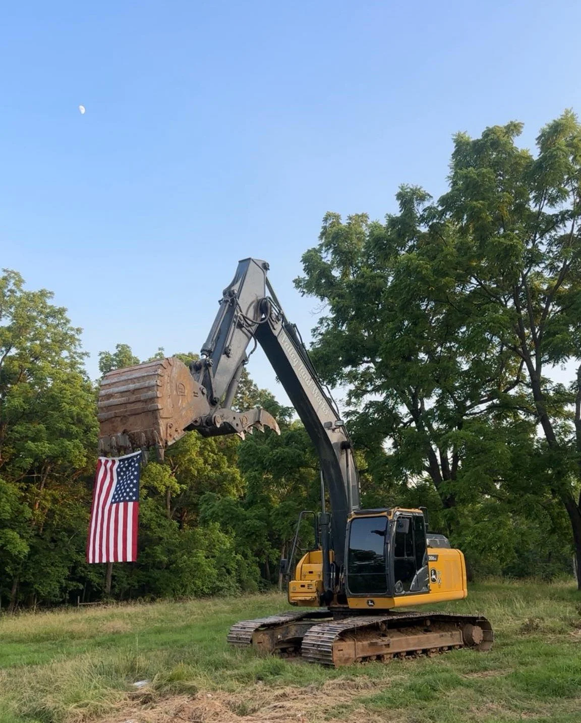 A yellow and black excavator holding an American flag attached to its bucket, with trees and a clear blue sky with a visible moon in the background.