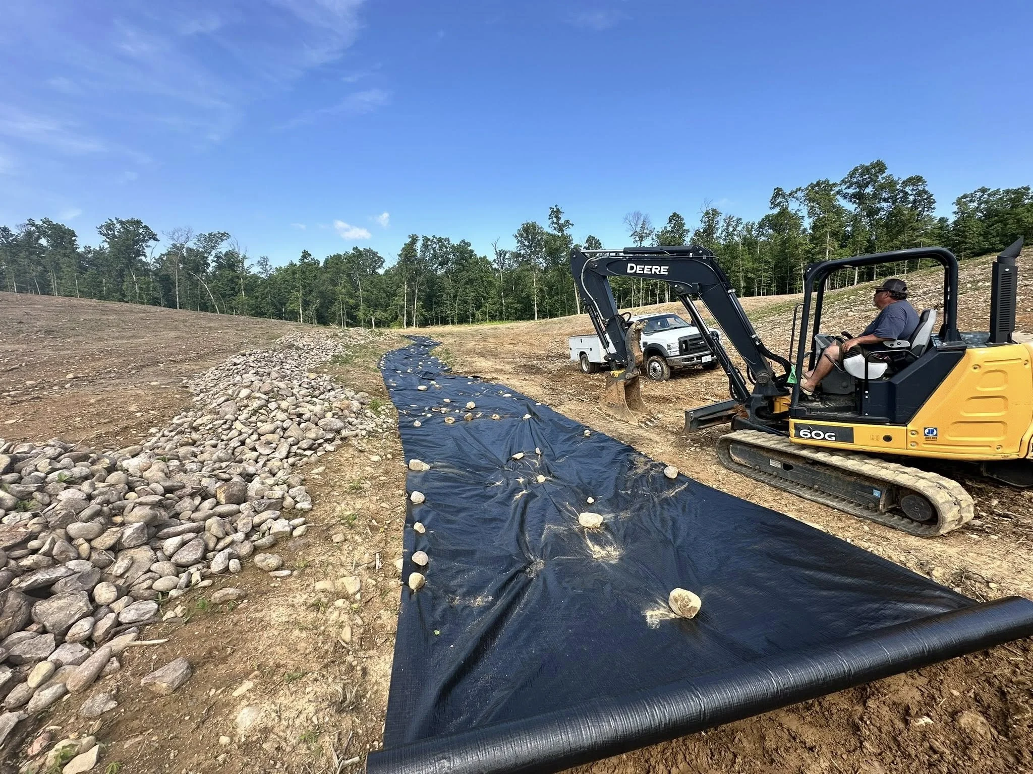 A construction site with a man operating a small excavator, a black plastic sheet on the ground, rocks lining the edge of a trench or pathway, and a forested area in the background under a blue sky.