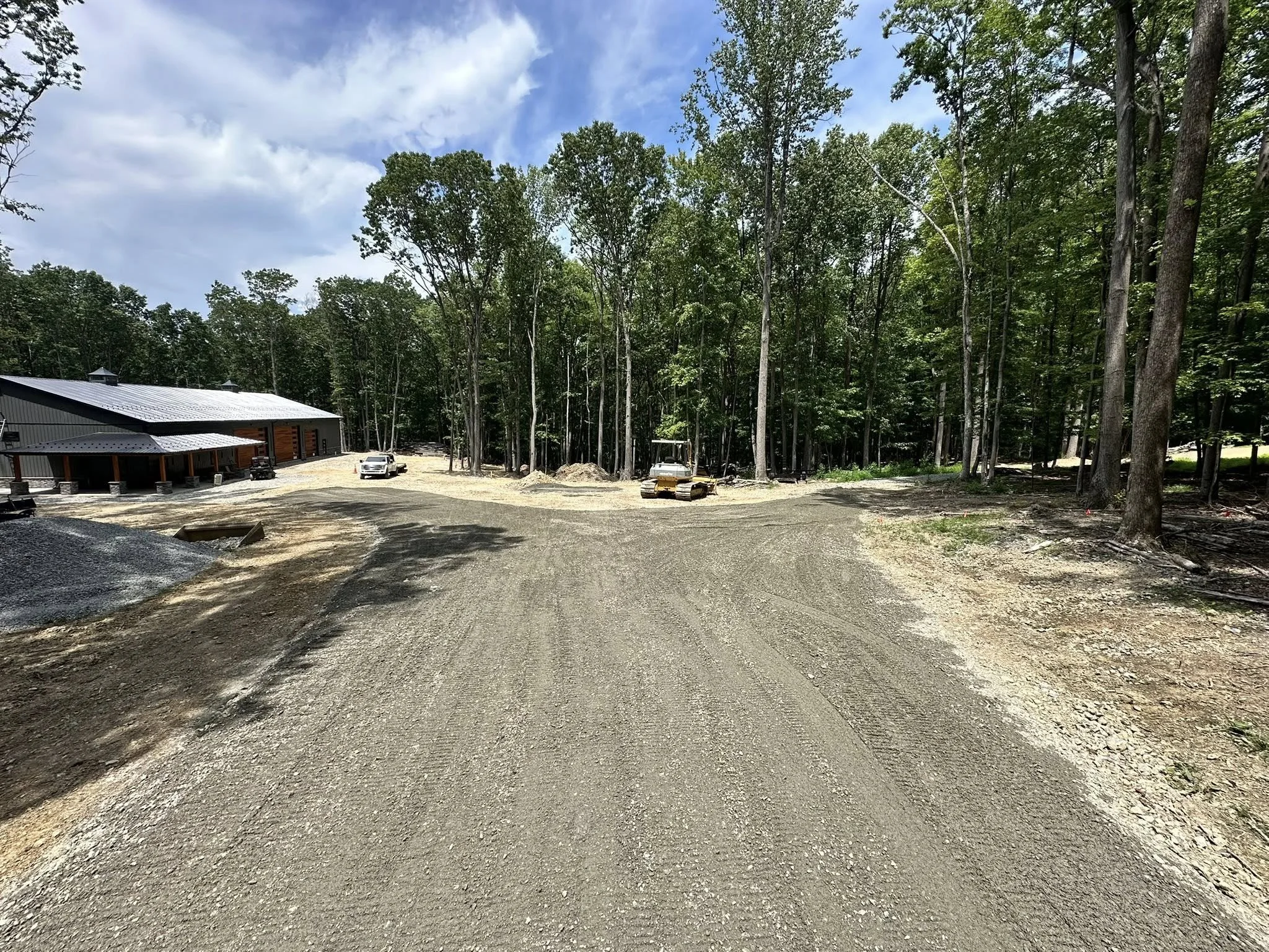 A construction site with a gravel road, a building with a metal roof, and a wooded area with tall trees in the background.
