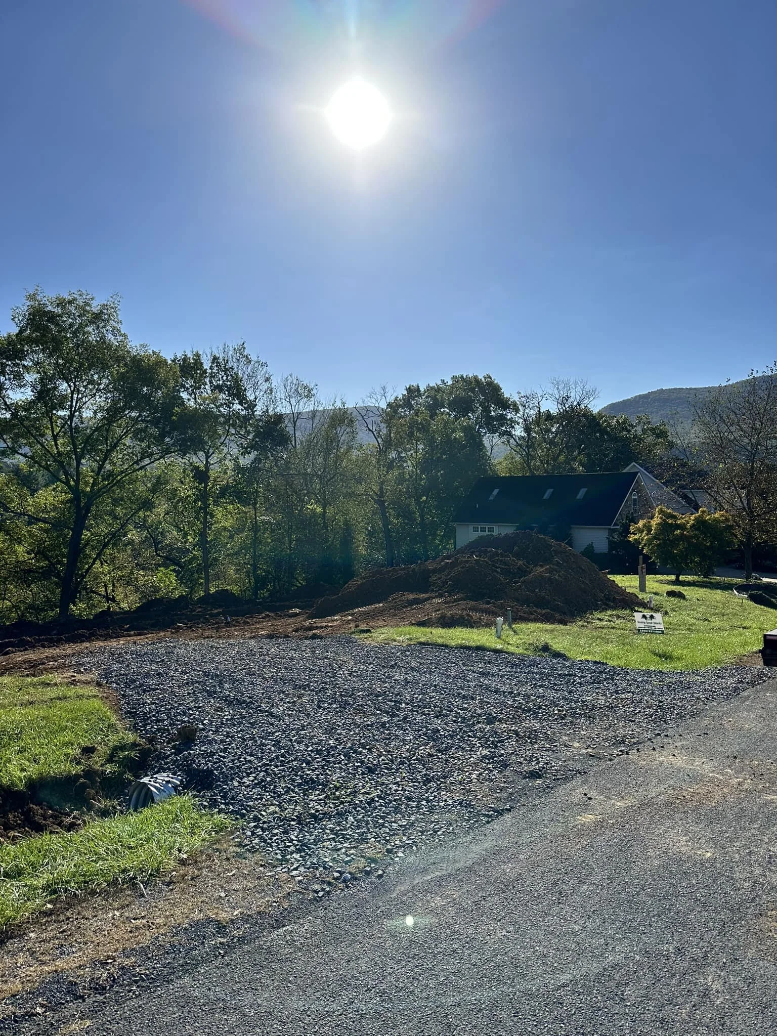A gravel pathway in a green area under a bright sun in a clear blue sky, with trees and a house in the background.