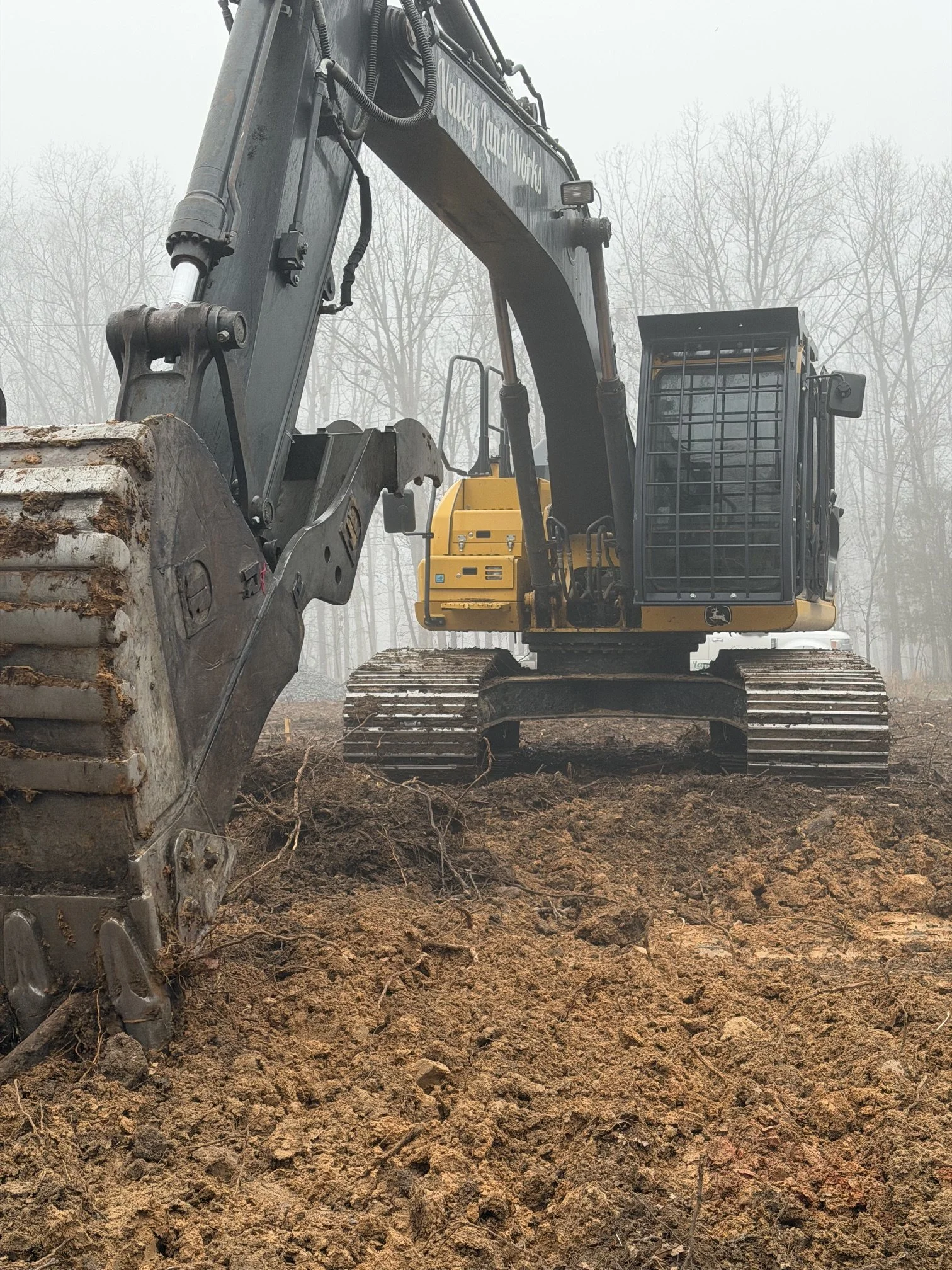A yellow and black excavator digging into soil in a foggy outdoor setting with leafless trees in the background.