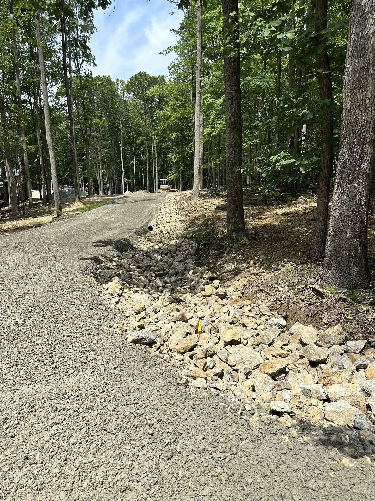 A dirt and gravel pathway through a forested area with trees on both sides, some construction equipment visible in the distance.