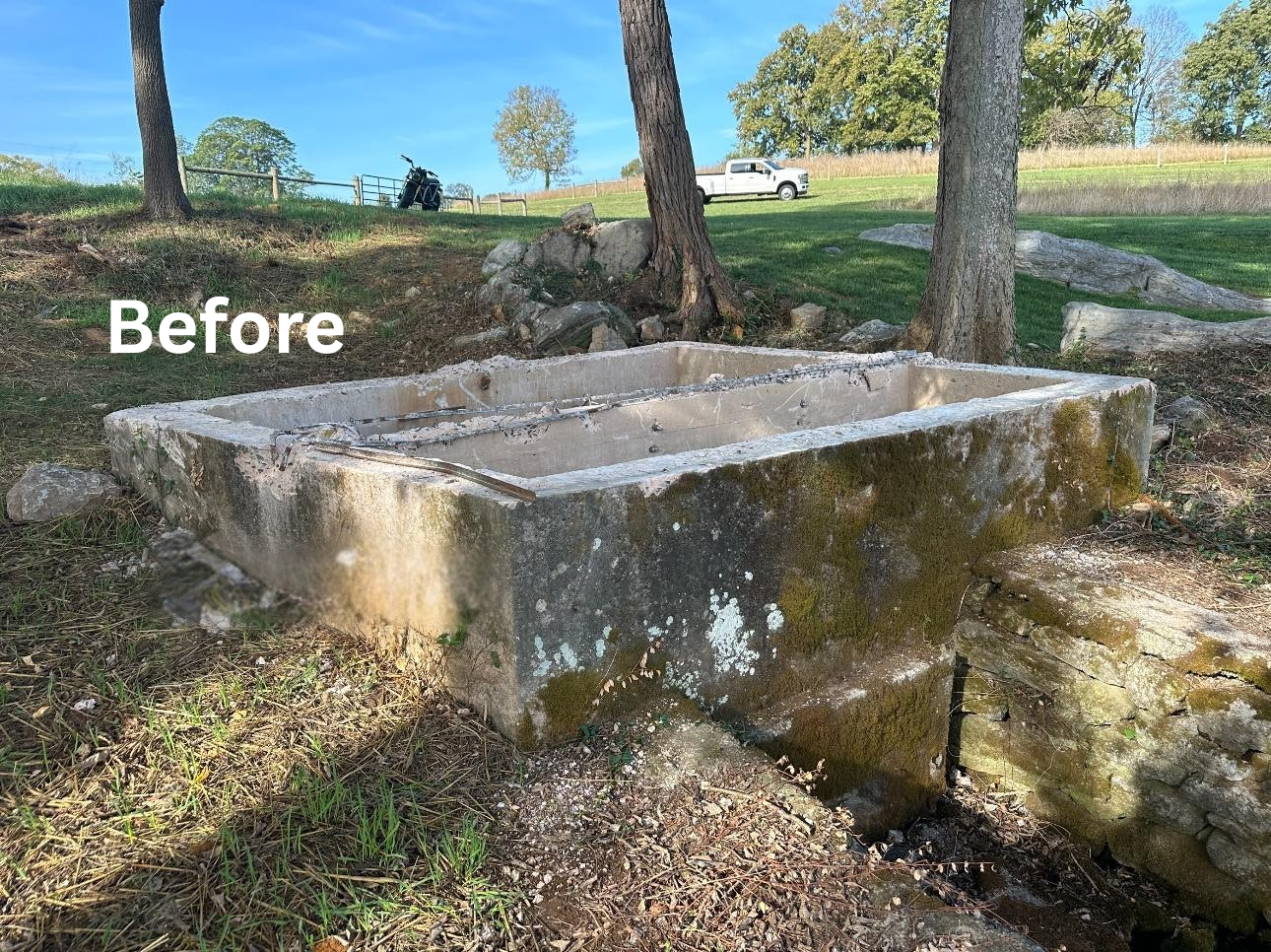 Old, weathered concrete water trough with moss and lichen, situated outdoors among trees, with a grassy area, rocks, a golf cart, and a pickup truck in the background.