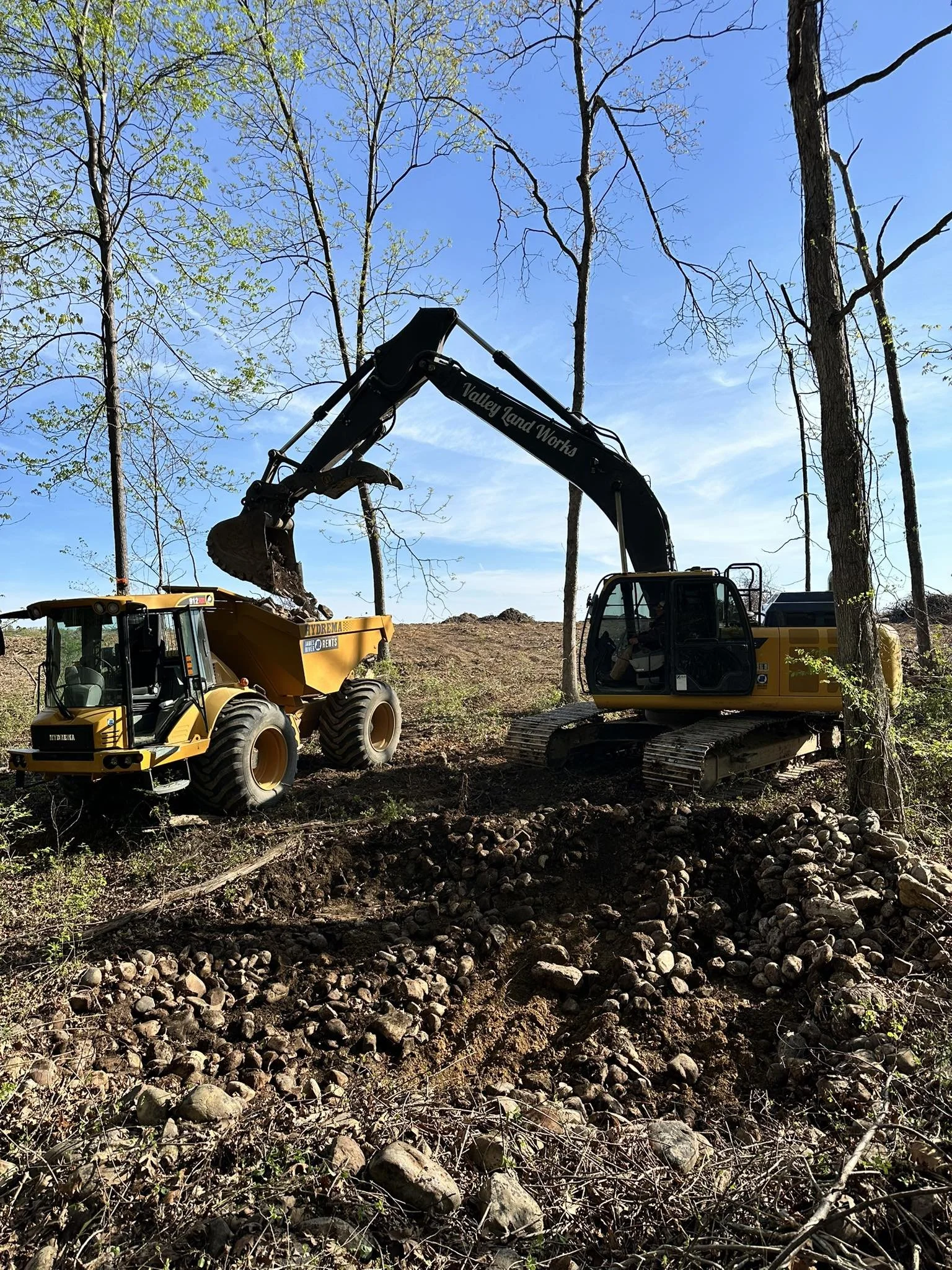 Construction equipment working on land with trees and a dirt, rocky surface under a clear blue sky.