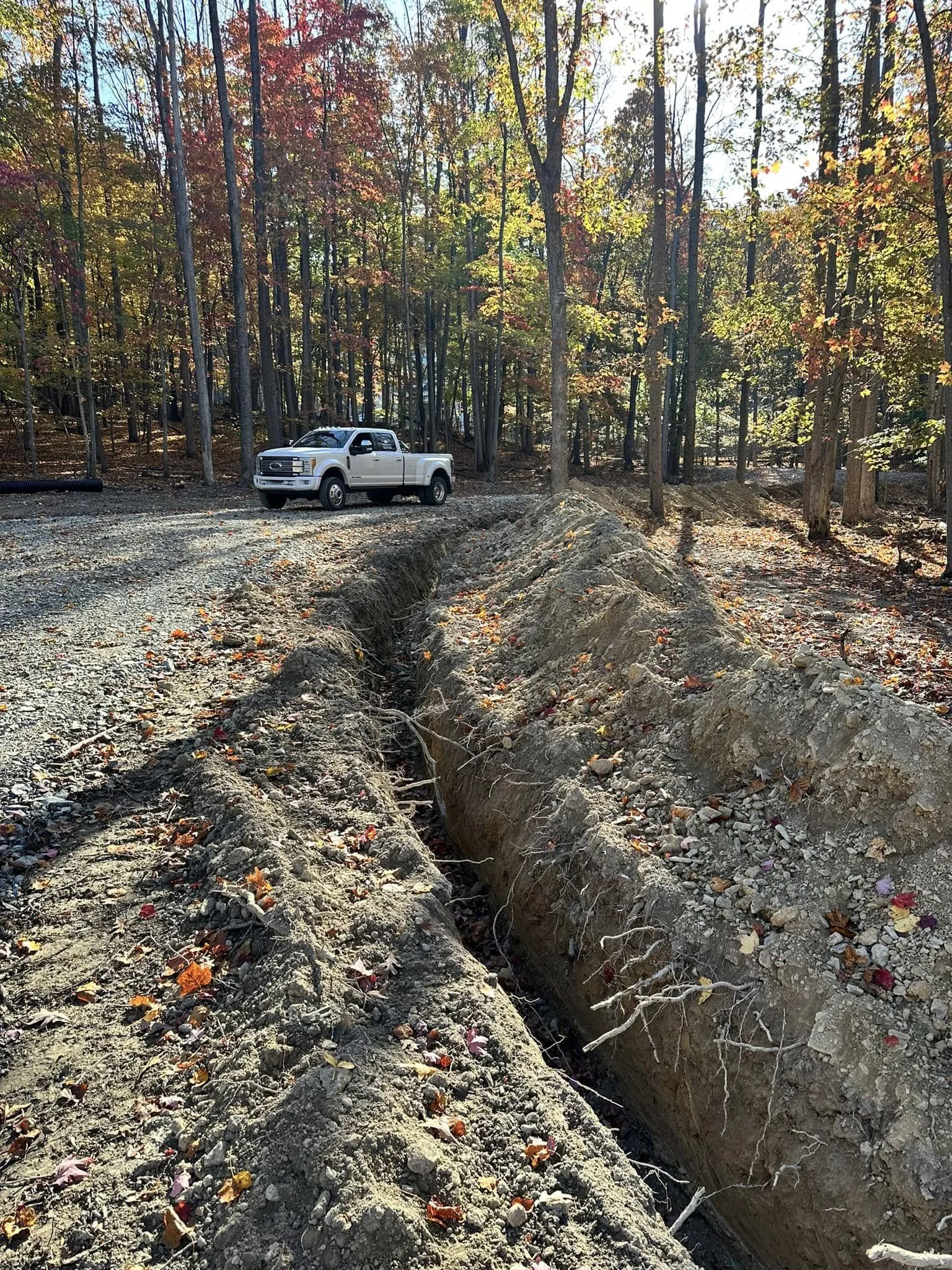 A large construction site in a wooded area with a deep trench, and a white pickup truck parked nearby on a gravel surface surrounded by trees with colorful fall foliage.