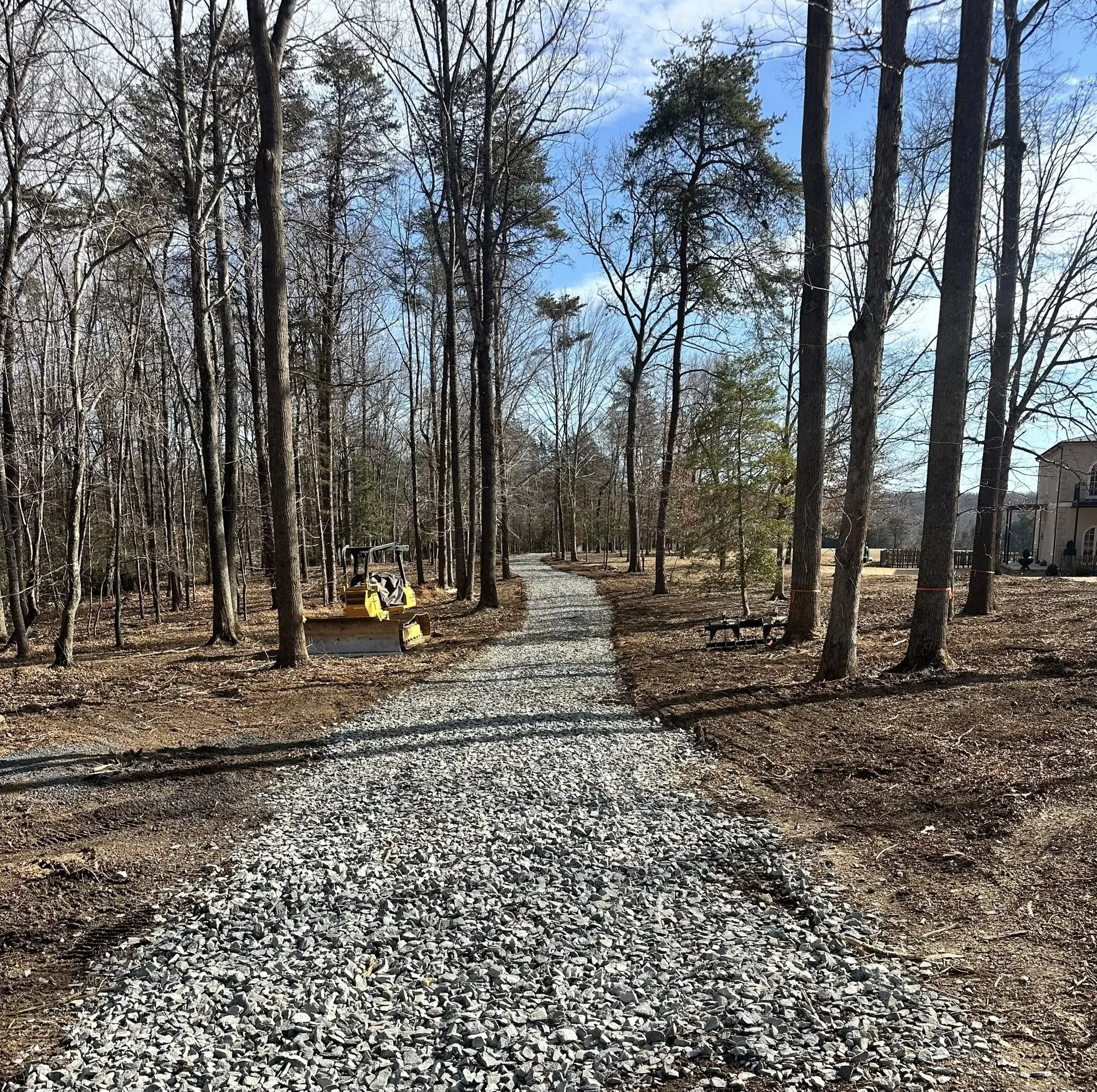 A gravel lane winding through a wooded area on a clear day with blue sky and some clouds. There is construction equipment on the left and a bench on the right.