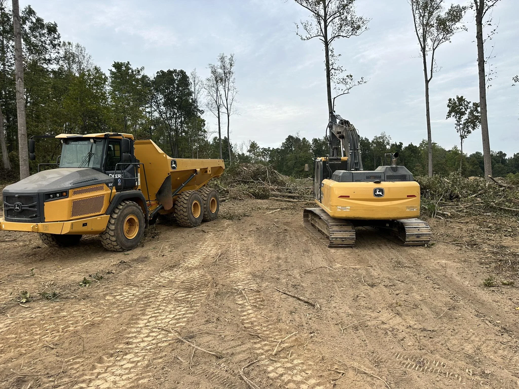 Construction site with a yellow dump truck and a yellow excavator on dirt ground, surrounded by trees in the background.